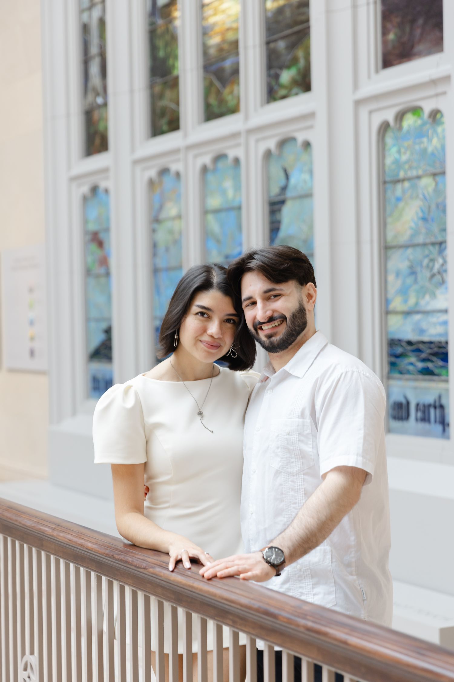 Couple in white attire posing together in front of colorful stained glass windows indoors.