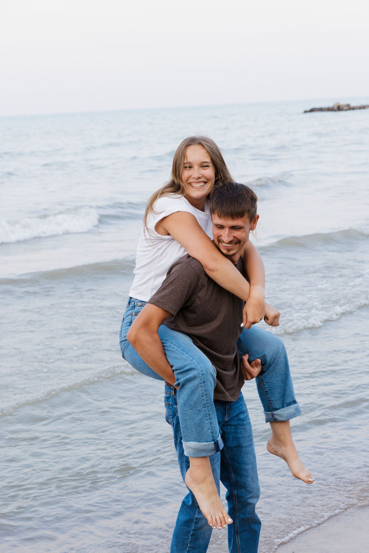 Happy couple enjoying a piggyback ride on the beach, smiling and wearing jeans and casual tops near ocean waves.
