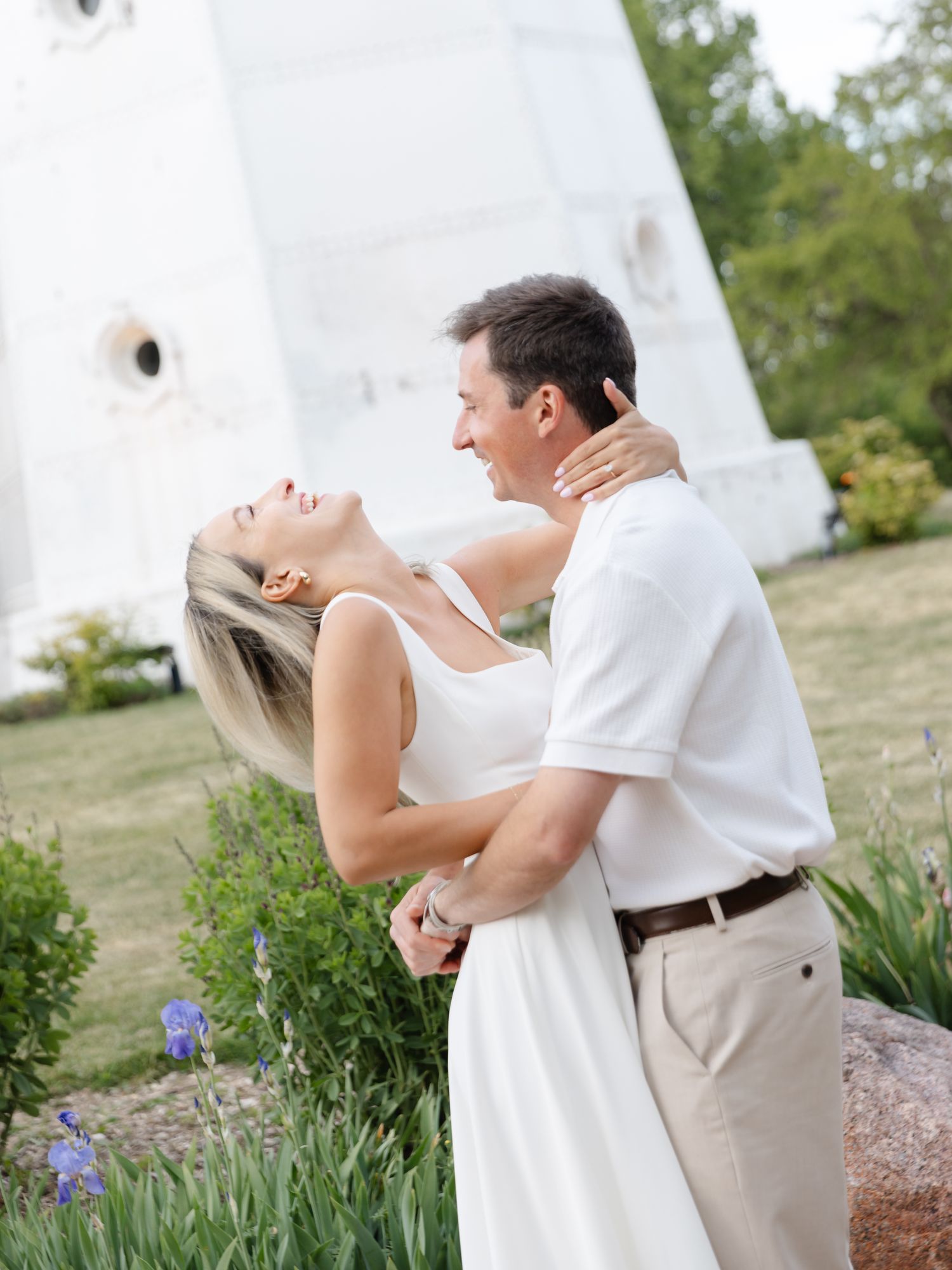 A couple embraces and laughs together in a garden near a white monument surrounded by blooming flowers.