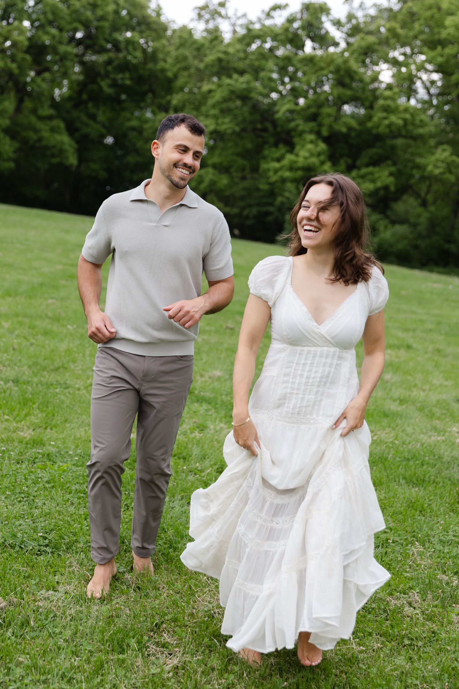 Happy couple walking barefoot on green grass, woman in white dress, man in gray.