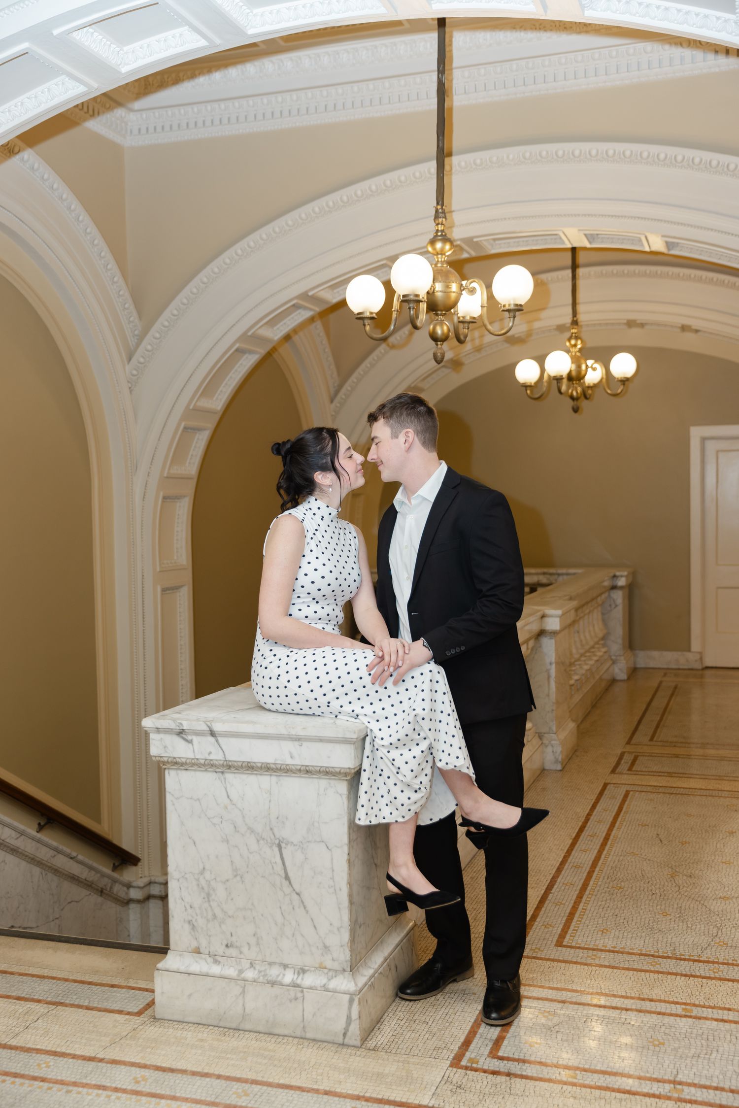 Elegant couple posing in ornate hallway with arched ceilings, woman in polka dot dress seated on marble pedestal.