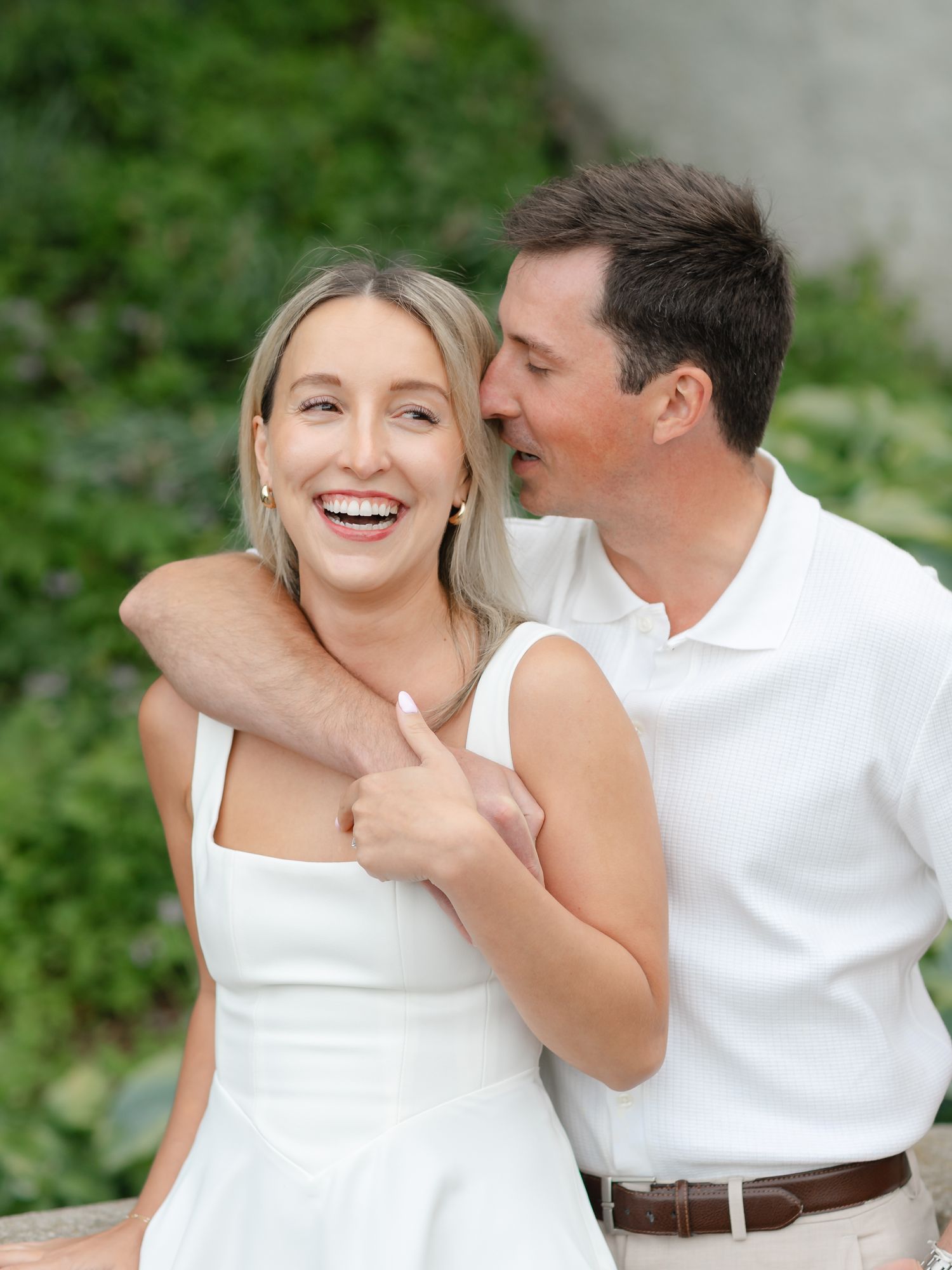 Happy couple in white outfits laughing together outdoors, man whispering to smiling blonde woman.
