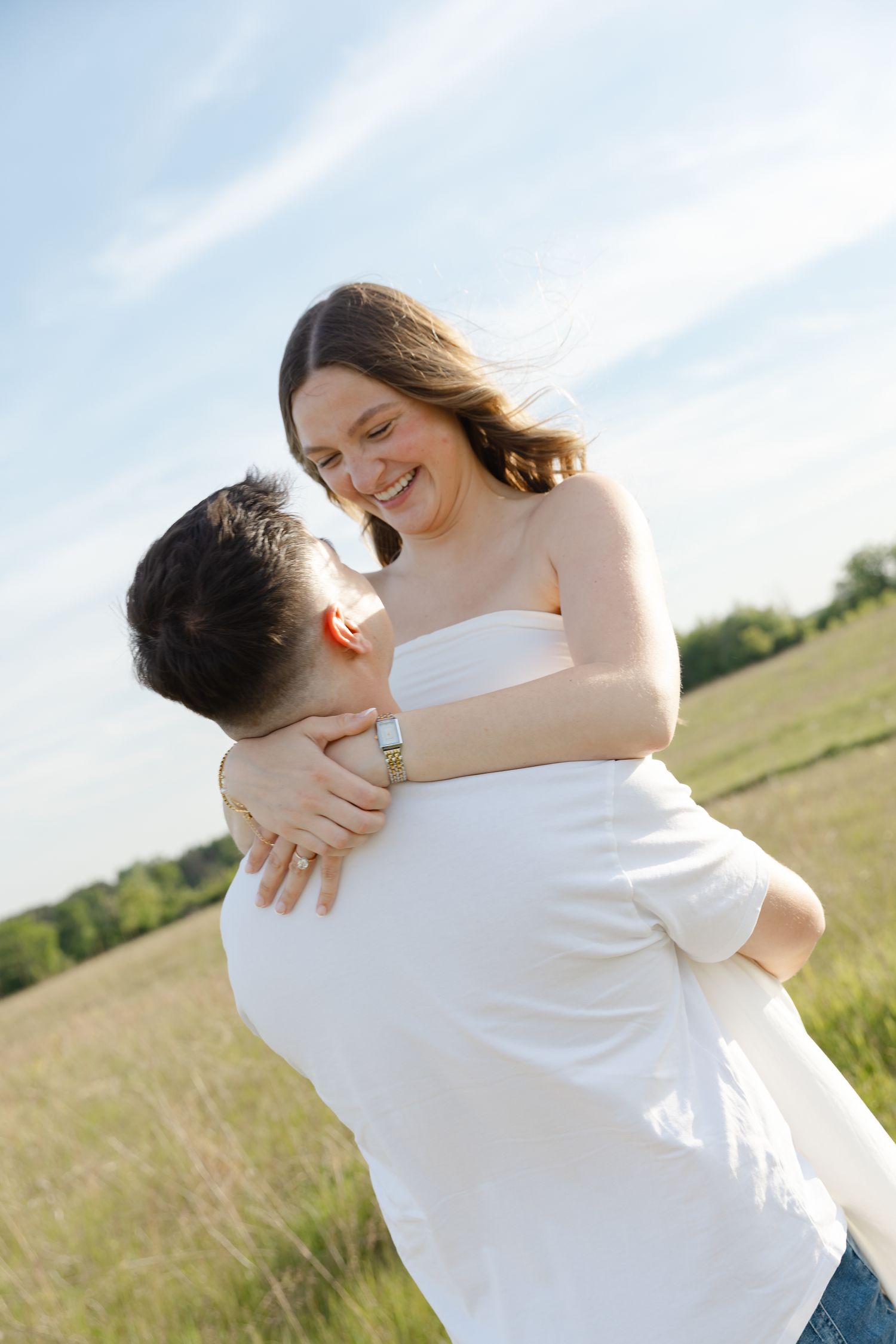 A happy couple embraces in a sunny green field, the man lifting the smiling woman in a white dress.