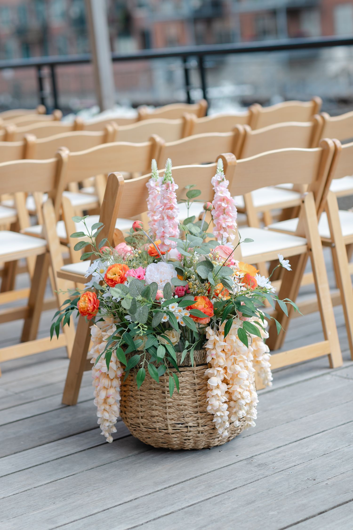Wicker basket filled with pink, orange, and white flowers on a wooden deck at an outdoor wedding ceremony.