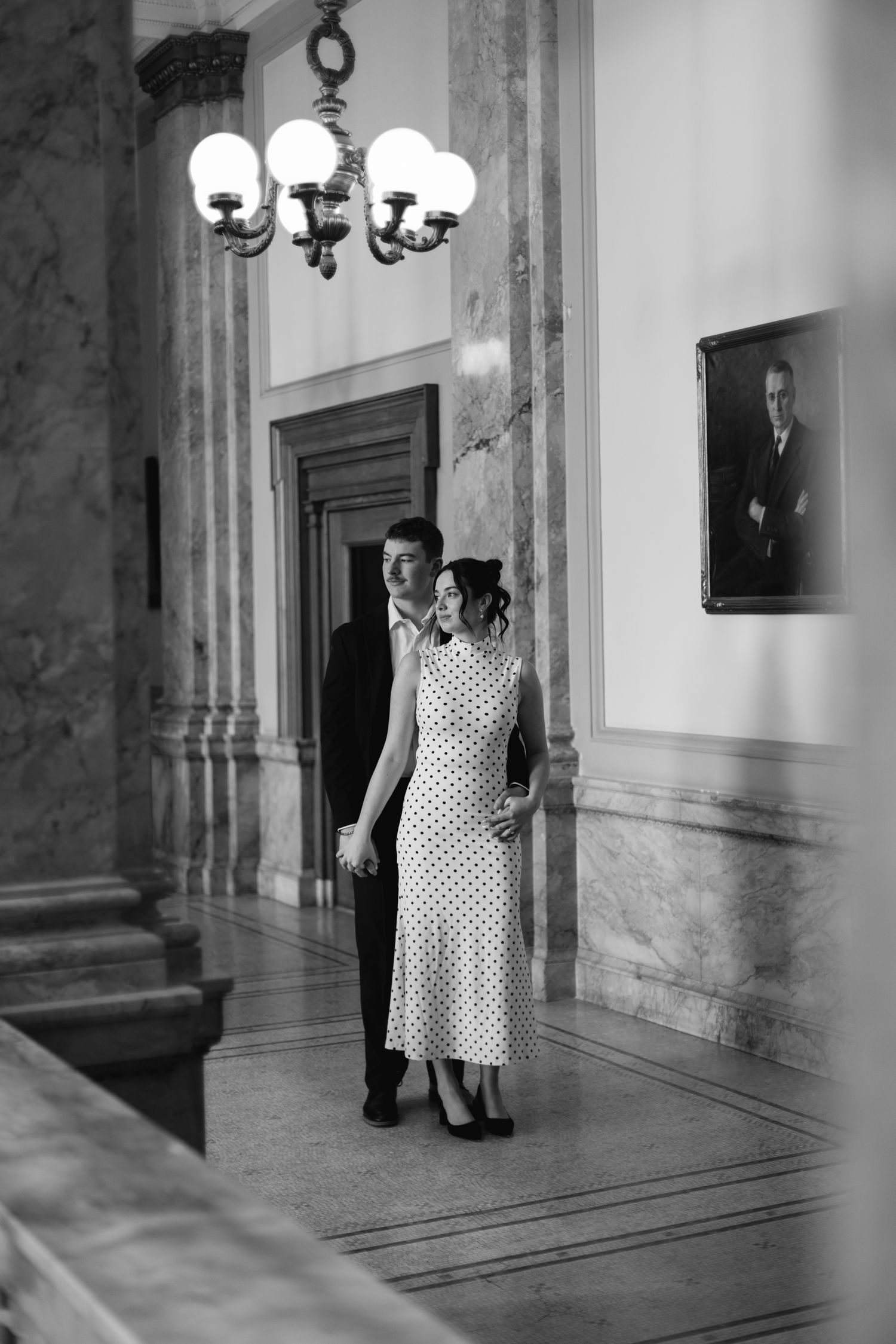 Black and white photo of couple walking through grand marble hallway with chandelier and portrait on wall.