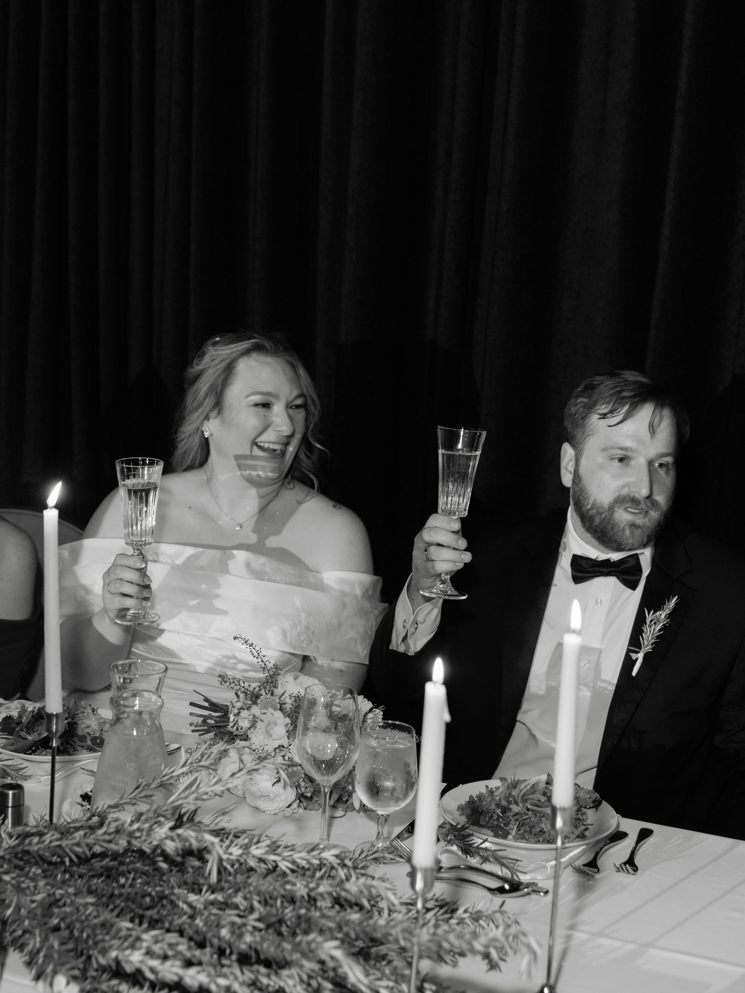 Black and white photo of a smiling bride and groom raising champagne glasses at their wedding reception dinner table.