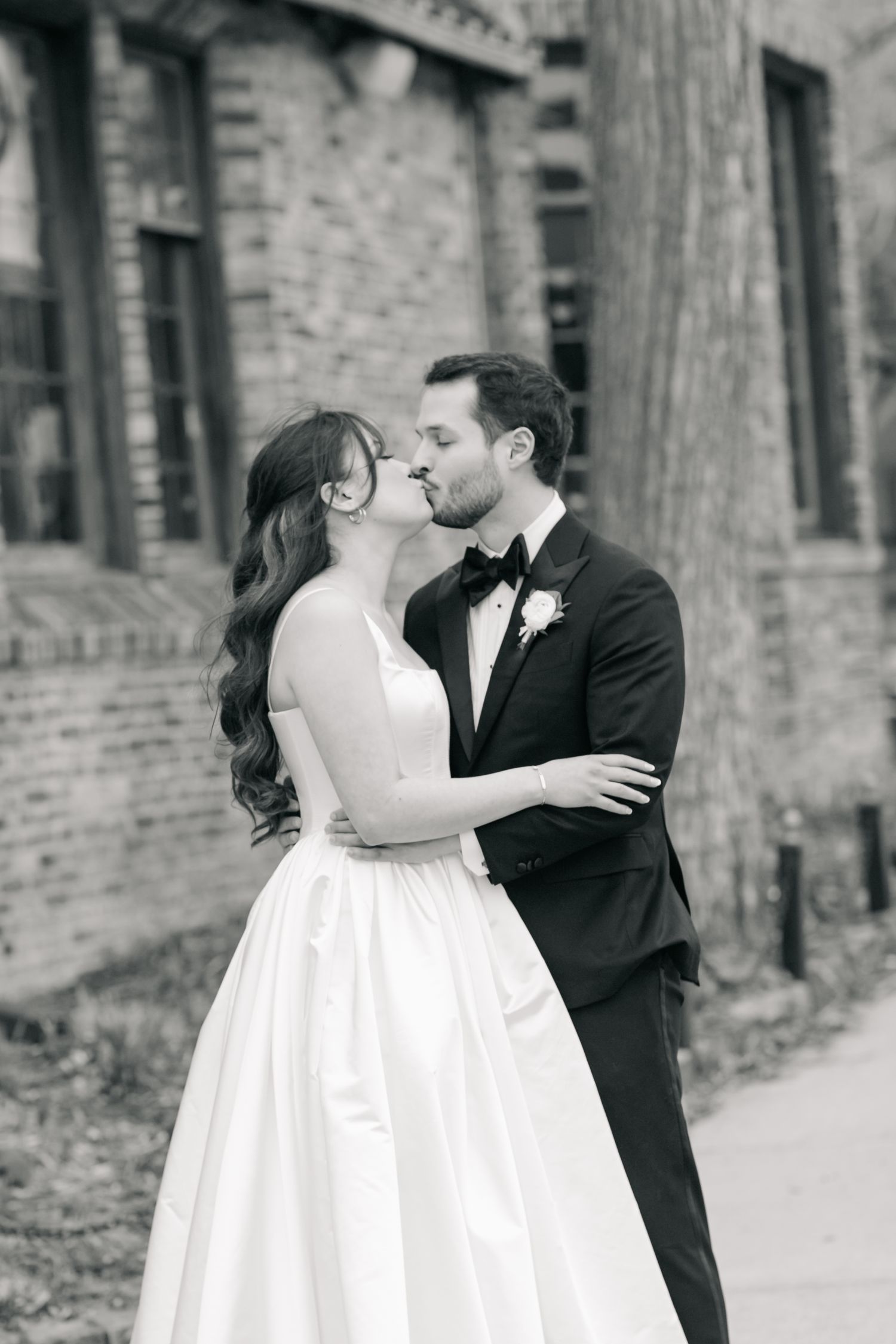 Black and white photo of a bride and groom sharing a kiss outside a brick building on their wedding day.
