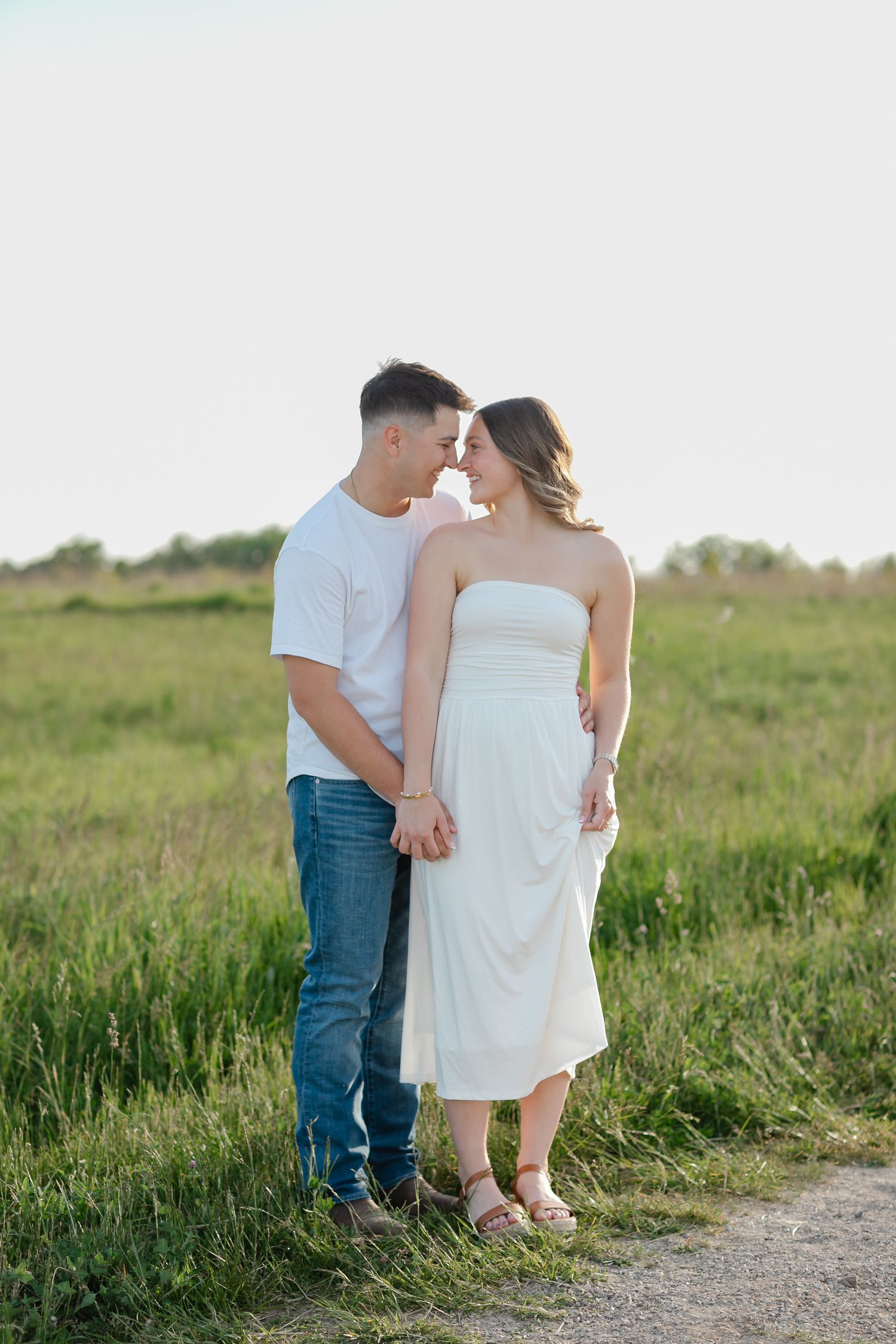 Couple in white dress and jeans holding hands in a green field, sharing a romantic moment outdoors.