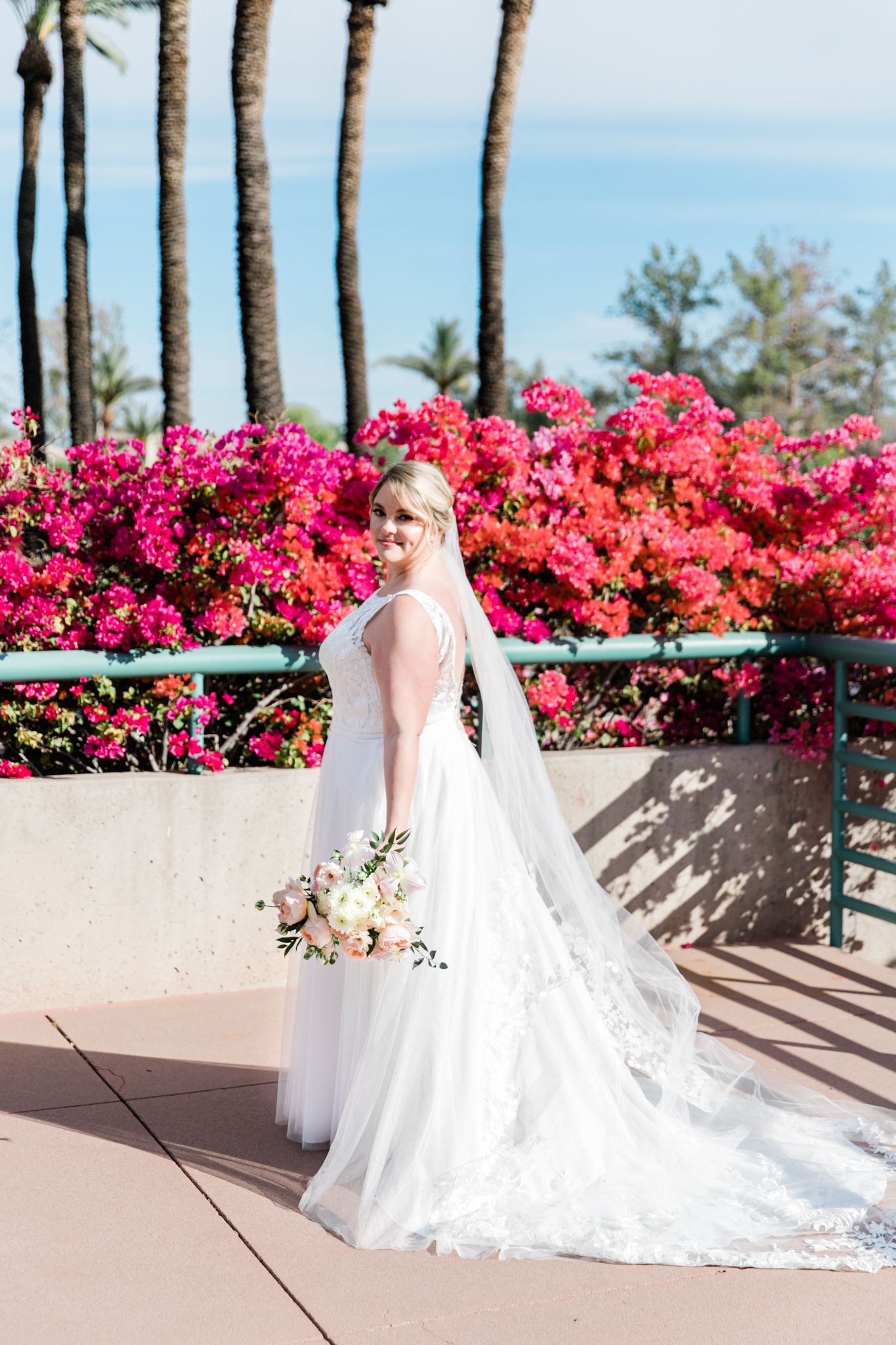 A bride in a flowing white gown poses by pink bougainvillea flowers against a coastal backdrop.