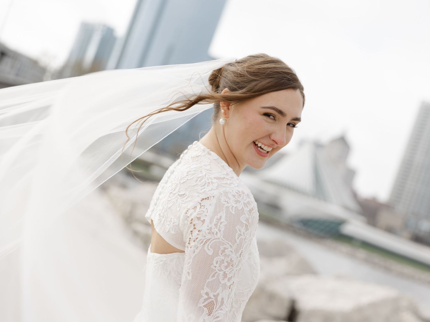 Smiling bride in lace wedding dress with veil blowing in wind, posing outdoors with city skyline background.