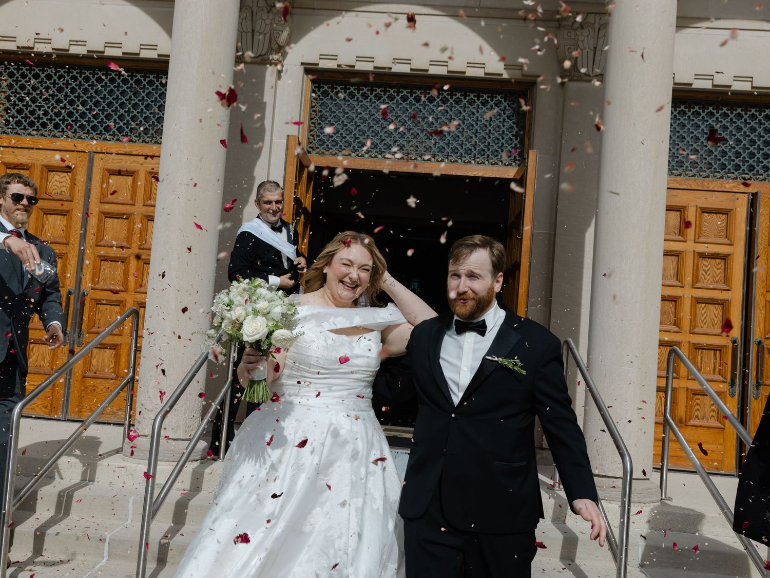 Joyful newlyweds exit church doors as guests toss colorful confetti, bride in white gown holding bouquet, groom in black suit.