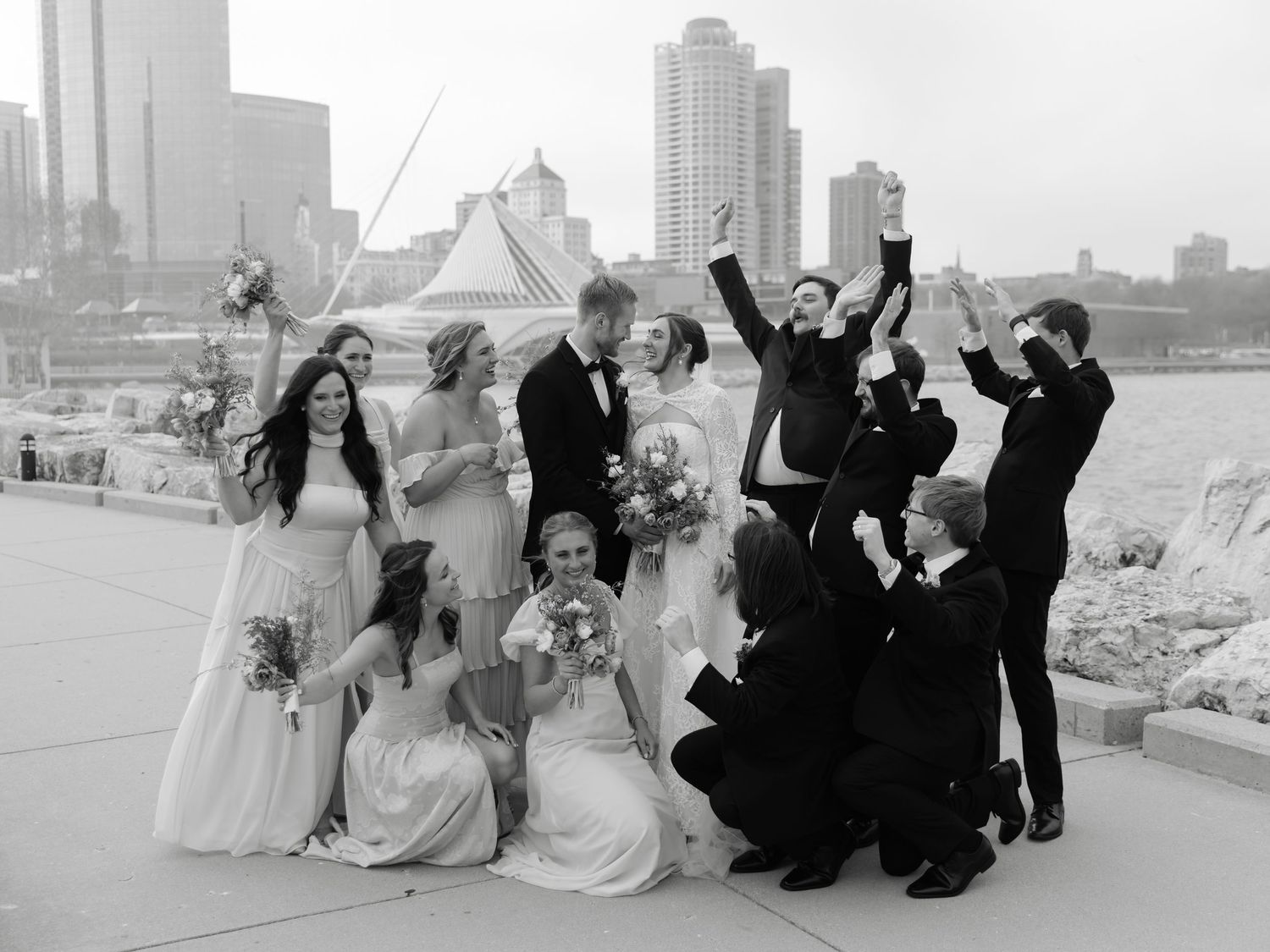 Black and white wedding party photo outdoors by waterfront with city skyline, celebrating joyfully.