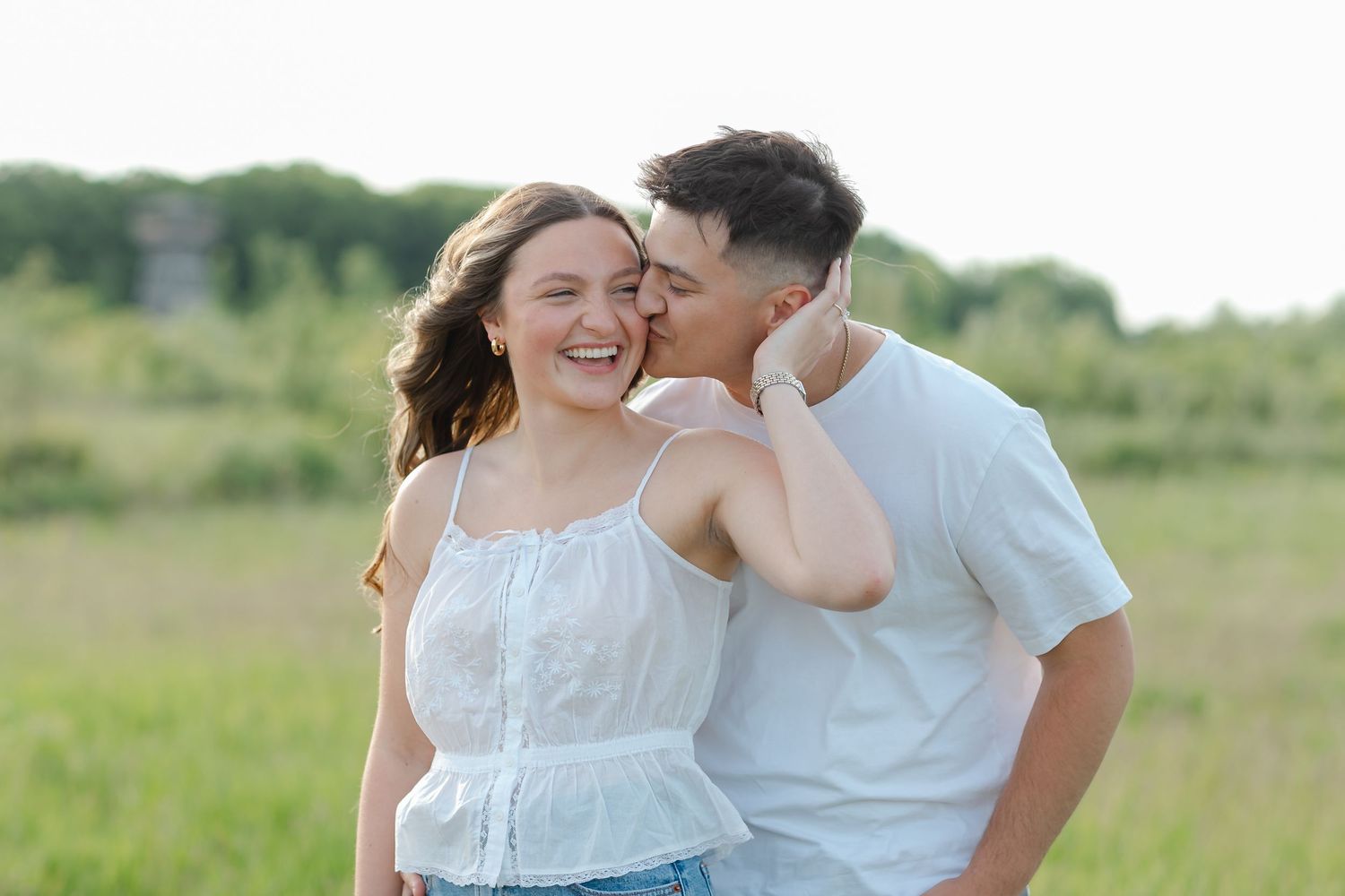 Young couple in white outfits sharing a tender moment in a green field, man kissing woman's cheek as she laughs.