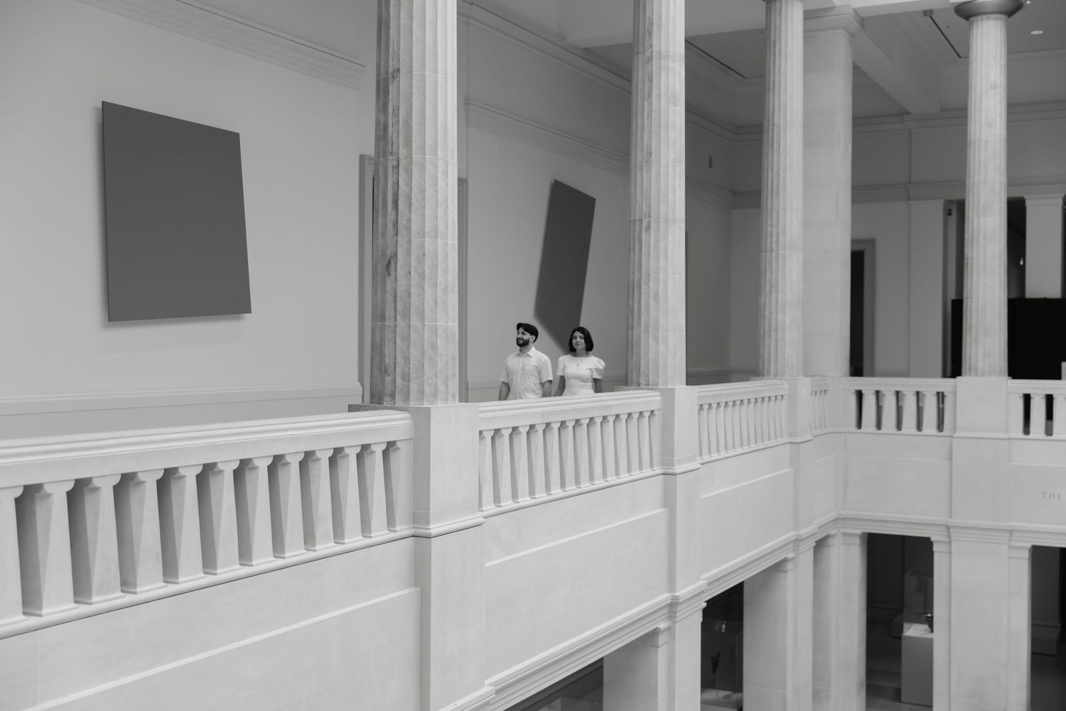 Two people stand on a white balcony in a classical museum gallery with large abstract paintings on the walls.