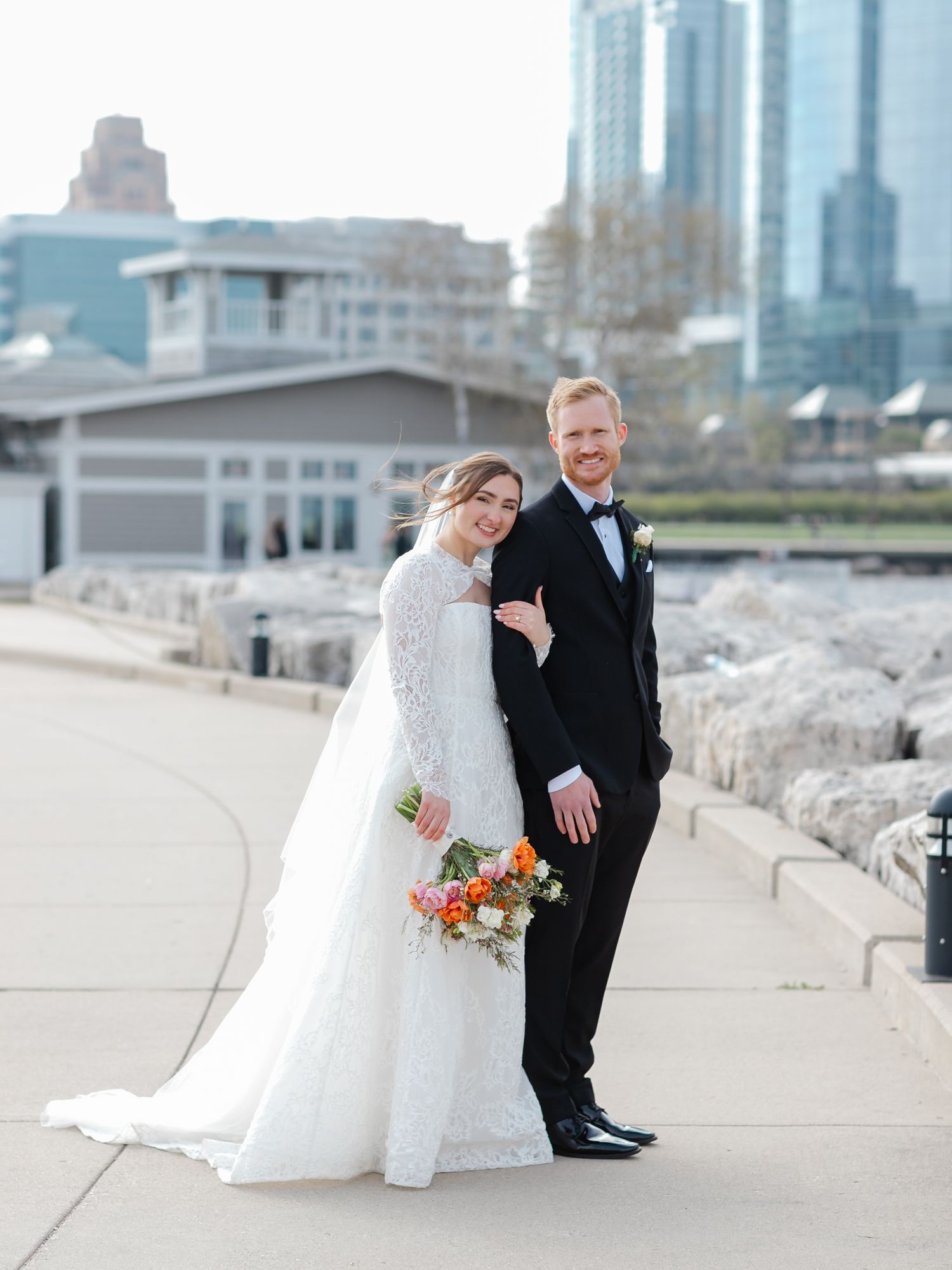 Bride in lace gown holds colorful bouquet beside groom in black tuxedo on lakeside path with city skyline.