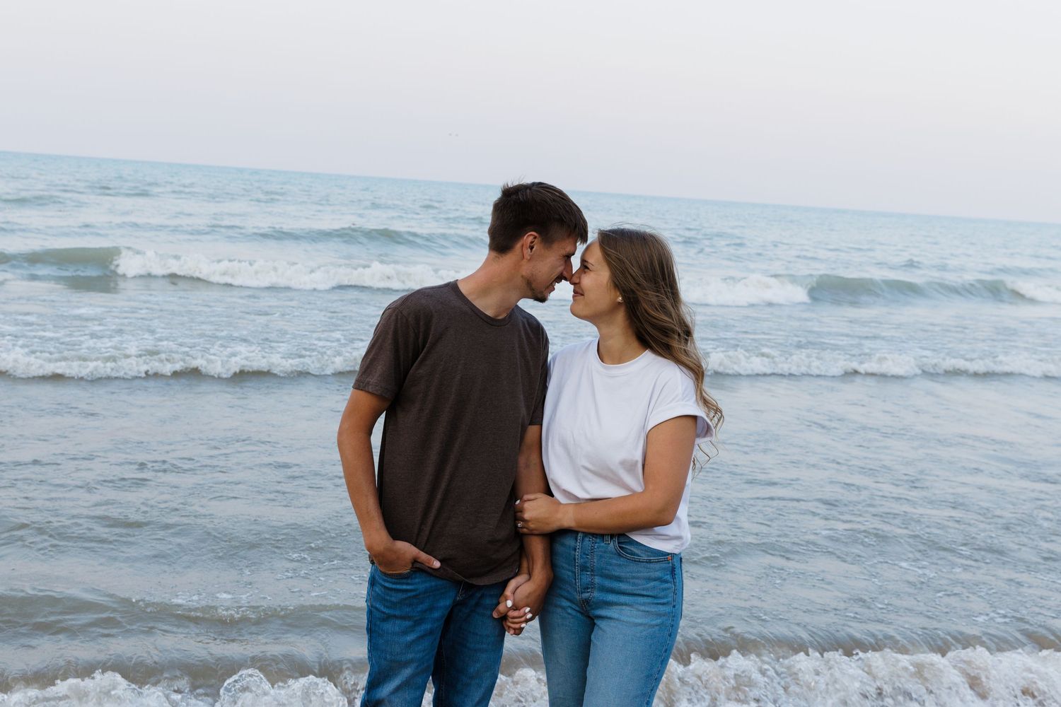 Romantic couple holding hands and touching foreheads on a beach with ocean waves in the background.