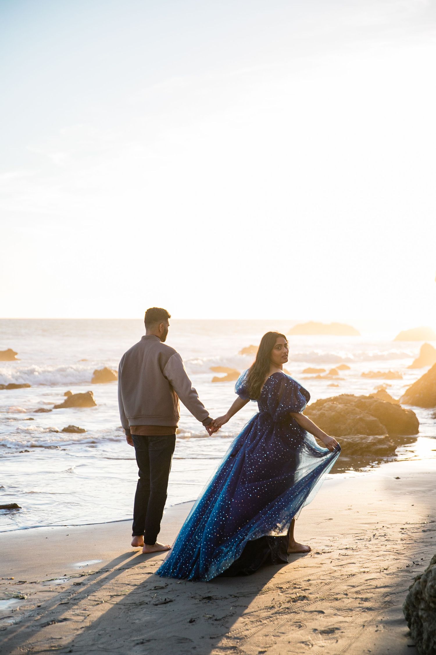 A couple sits on a beach at sunset holding hands while waves crash against rocks in the background.