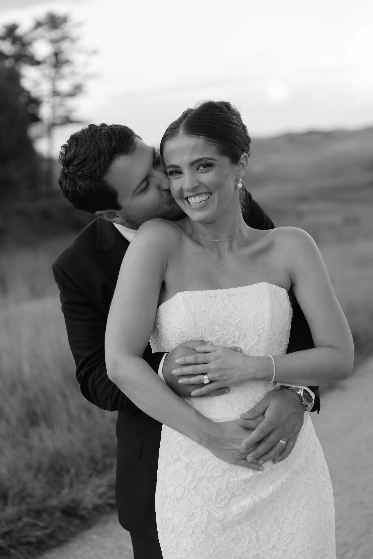 Black and white photo of a groom kissing his bride on the cheek while embracing her outdoors at sunset.
