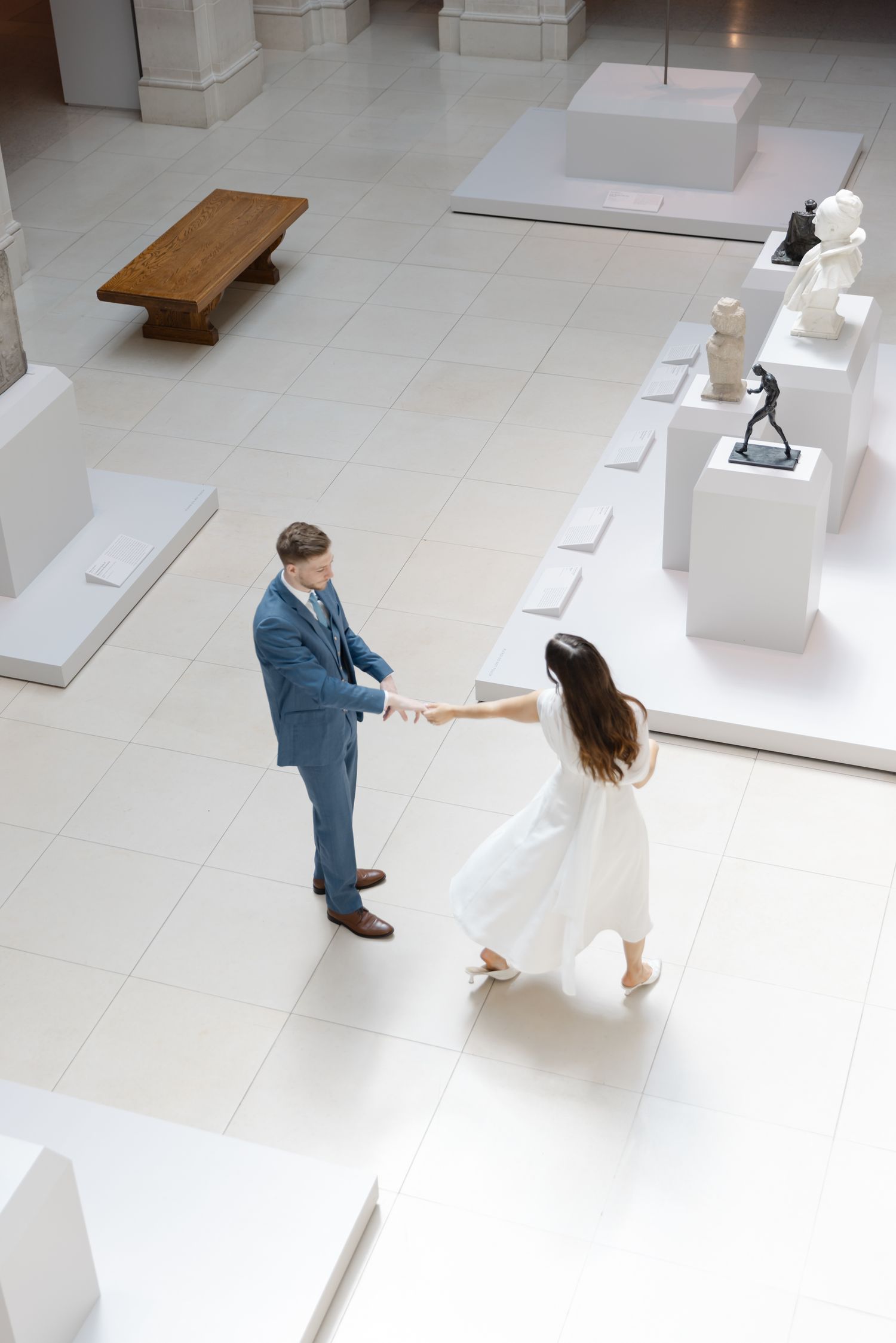Couple in wedding attire holding hands and dancing in a bright museum gallery with white pedestals and sculptures.