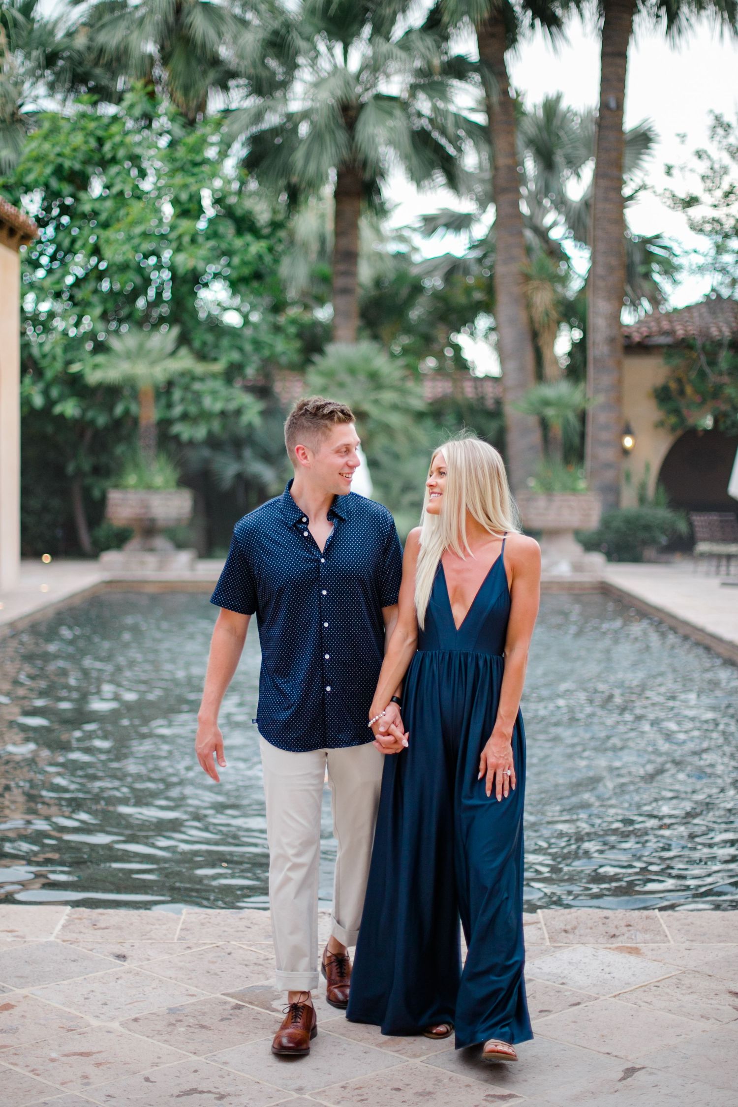 A couple in matching navy blue outfits walks along a palm tree-lined waterfront during a romantic photoshoot.