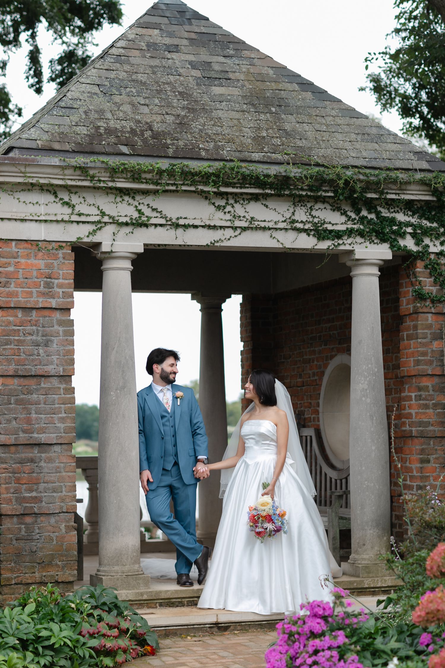 Bride and groom holding hands at a brick gazebo with stone columns, surrounded by lush gardens and colorful flowers.