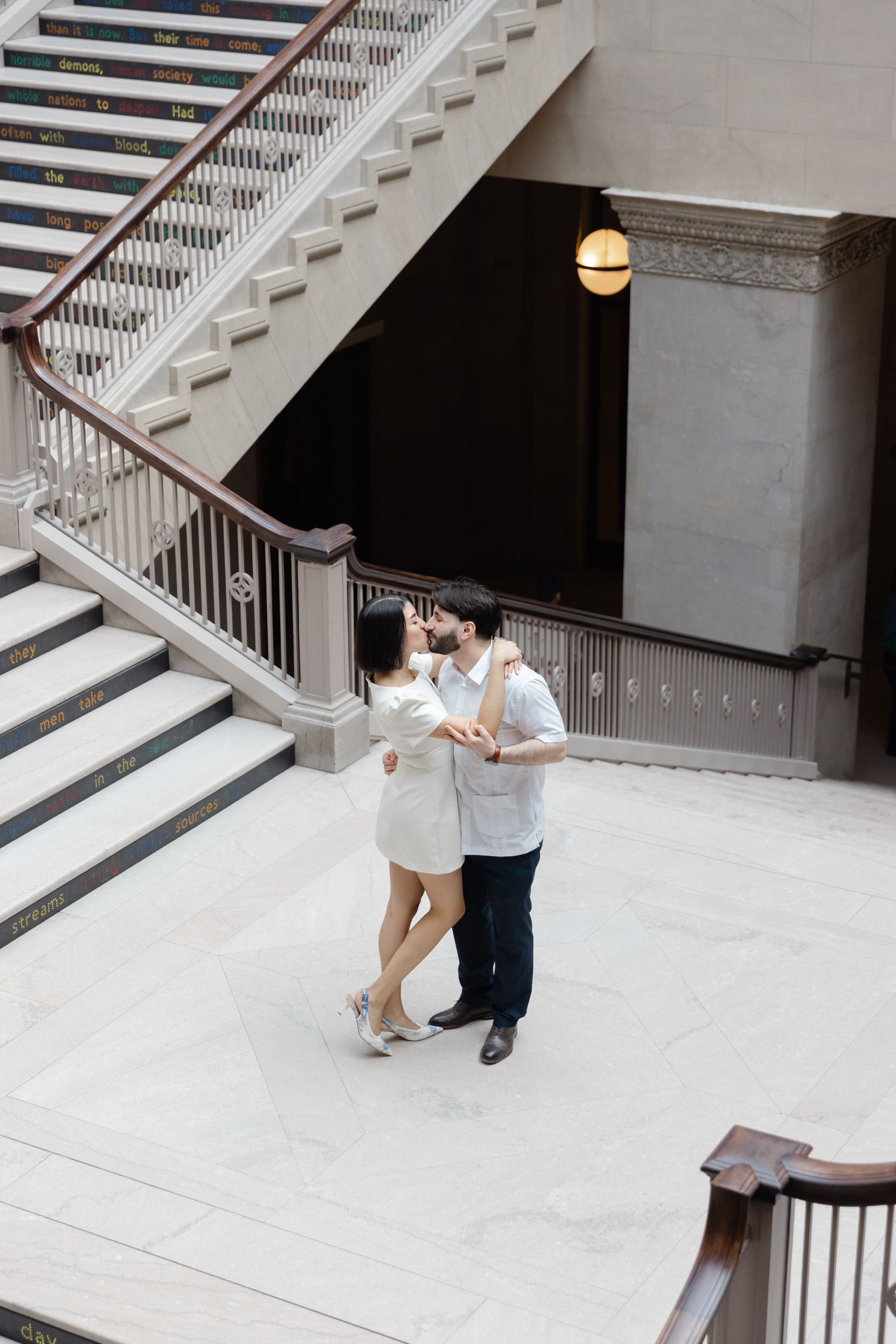 Romantic couple embracing and kissing at the base of a grand marble staircase in a museum.