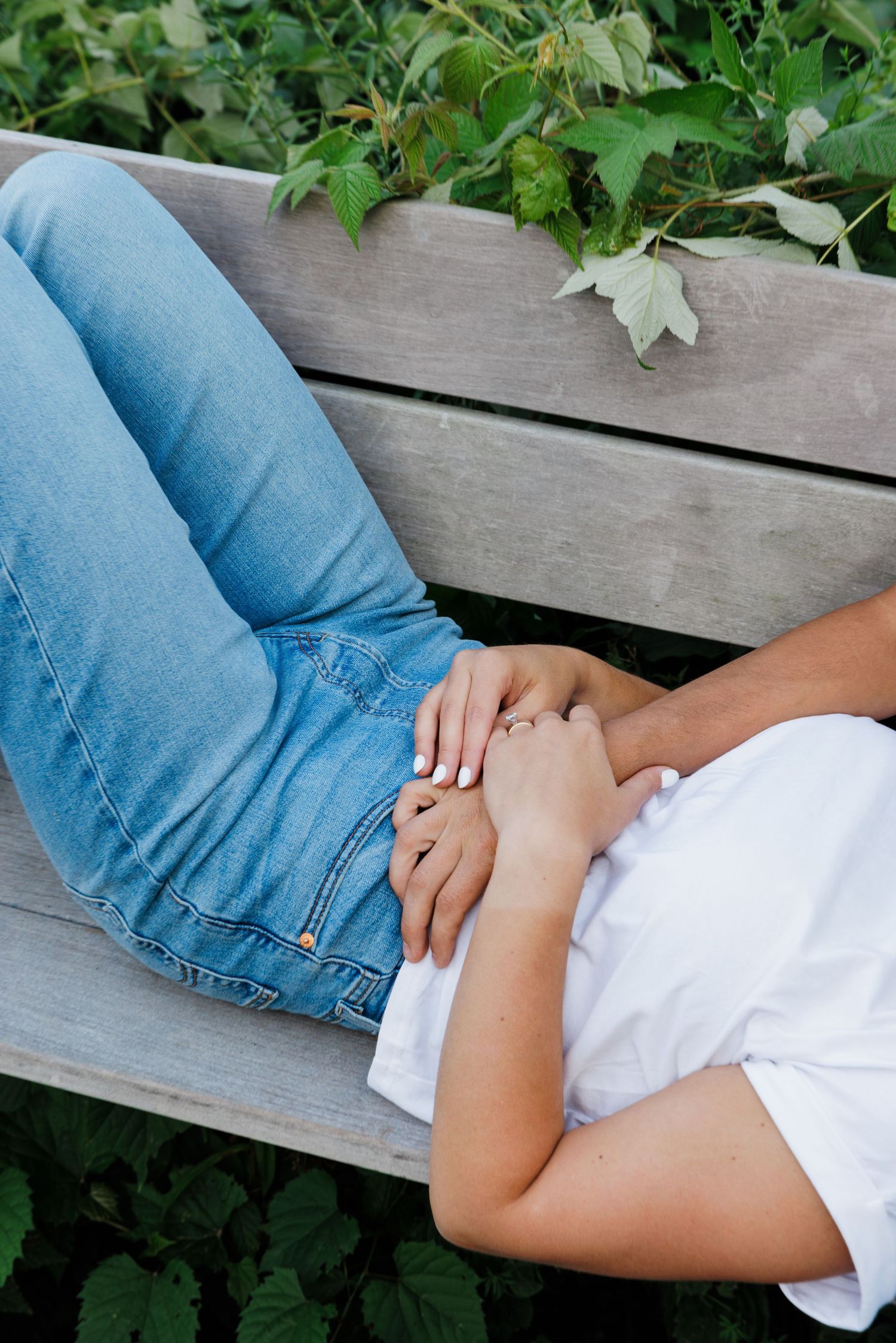 Couple holding hands while relaxing on a wooden bench surrounded by green plants outdoors.