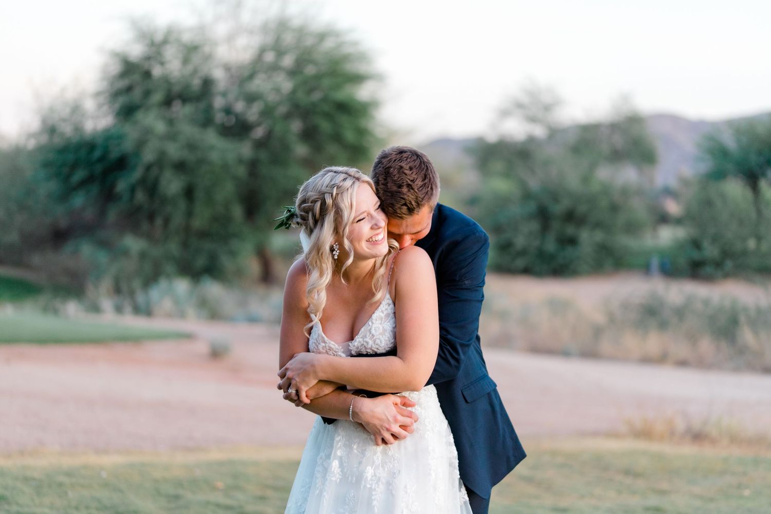 A couple embraces outdoors in a desert landscape with mountains and trees in the background.