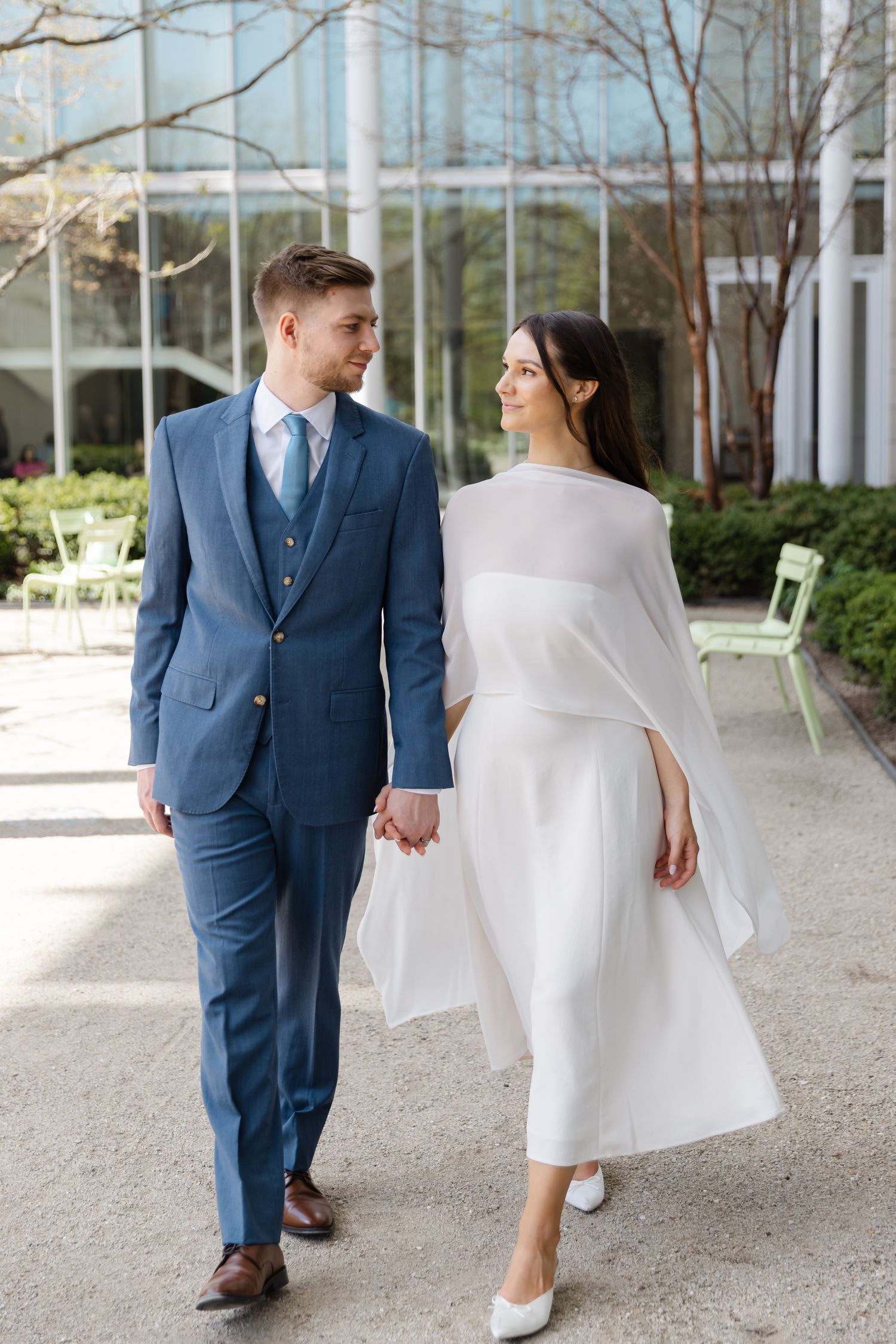 Couple in wedding attire holding hands outdoors, groom in blue suit and bride in white midi dress.