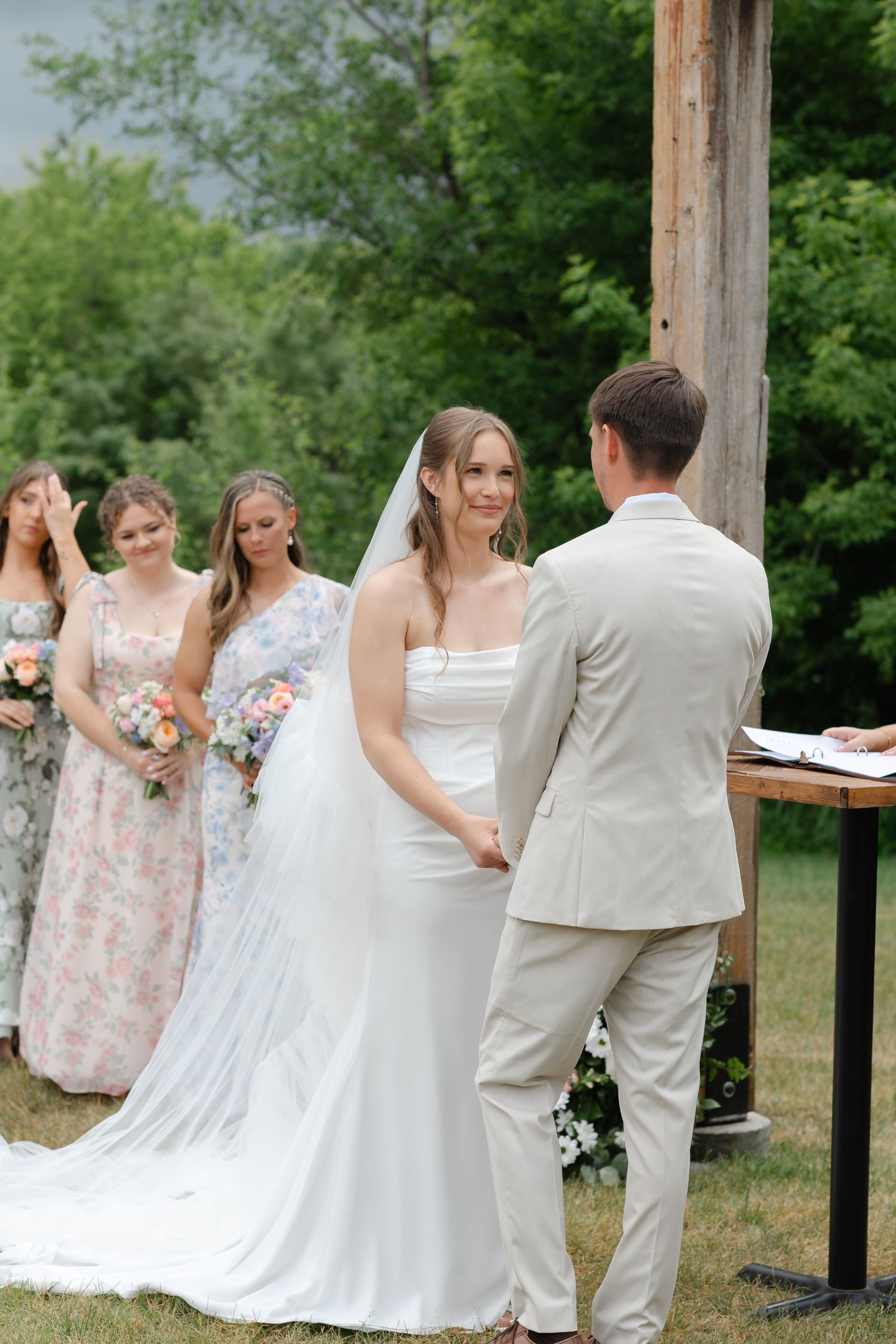 Bride and groom exchange vows at outdoor wedding ceremony with bridesmaids in floral dresses holding bouquets.