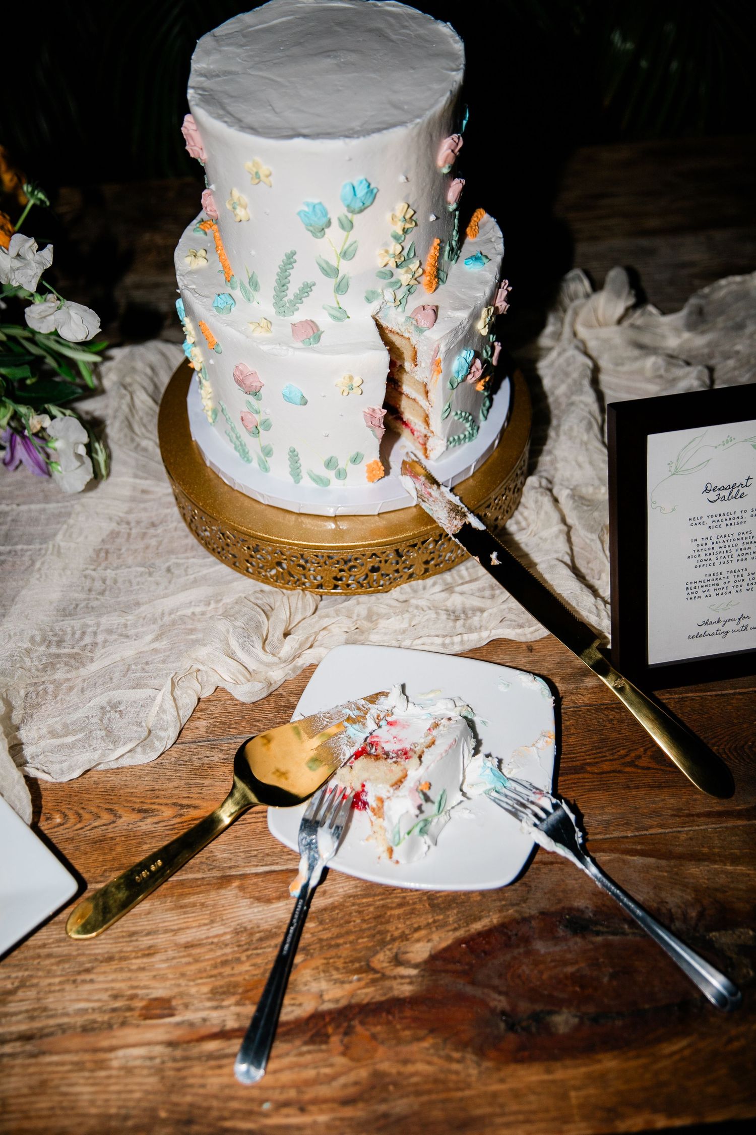 A white wedding cake with pastel floral decorations sits on a gold stand with serving utensils.