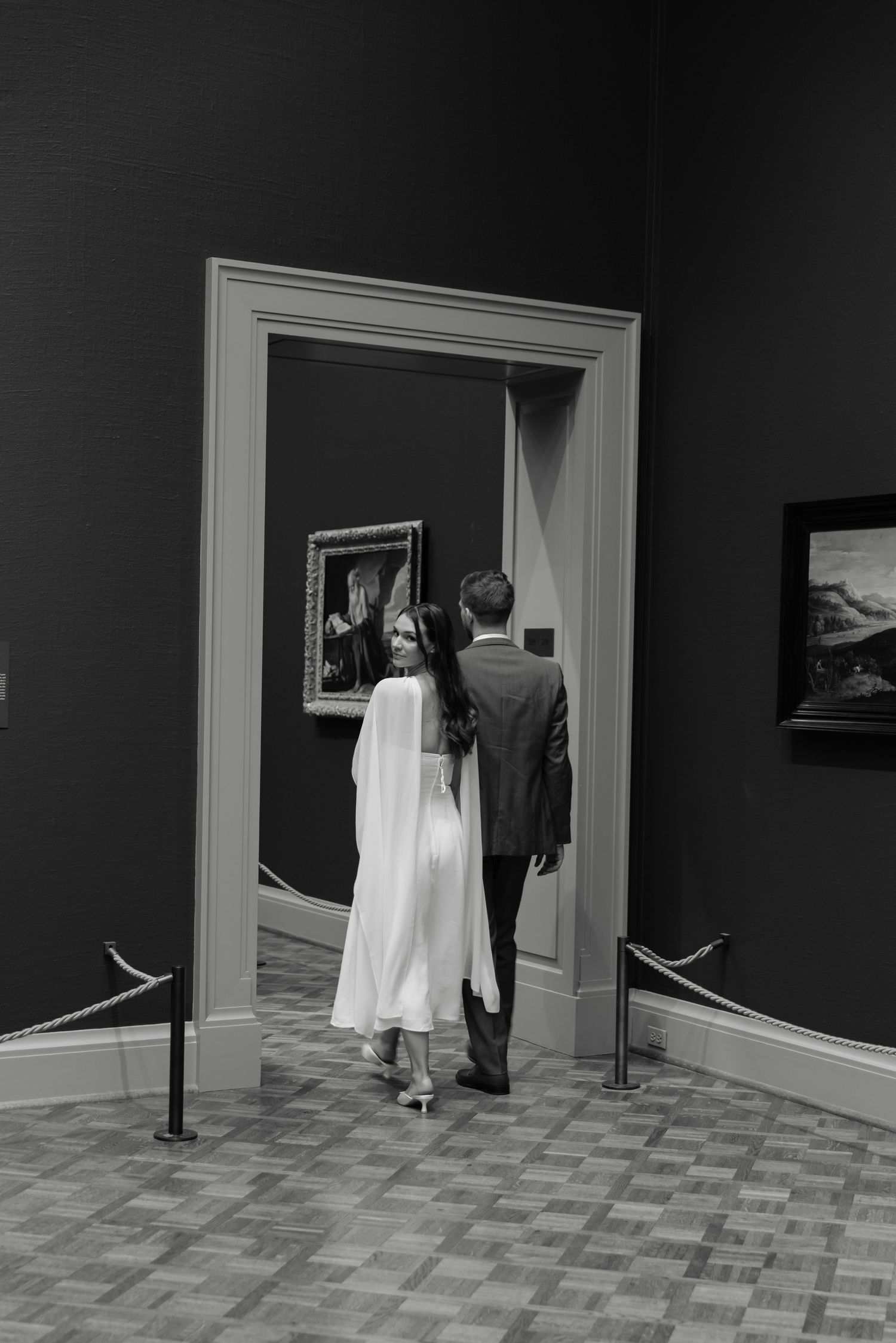 Black and white photo of a couple in formal wear walking through an art museum gallery doorway.