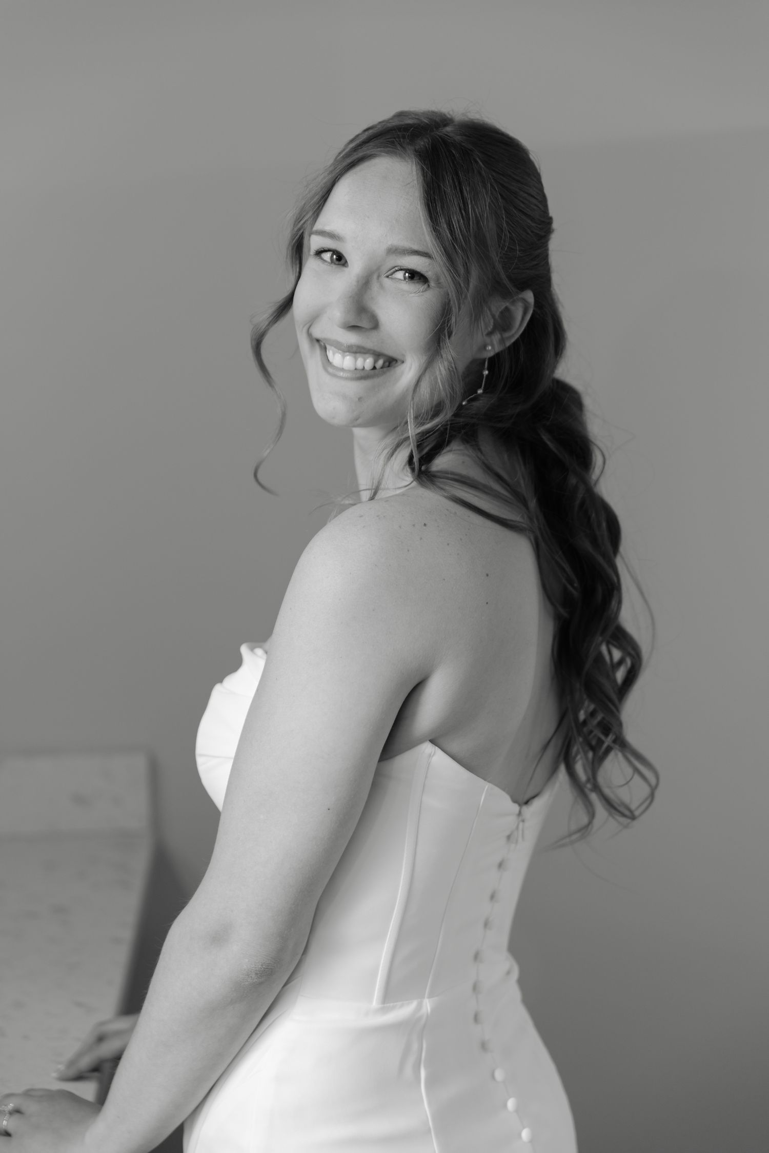 Black and white portrait of a smiling bride in a strapless gown with button back, hair in loose curls.