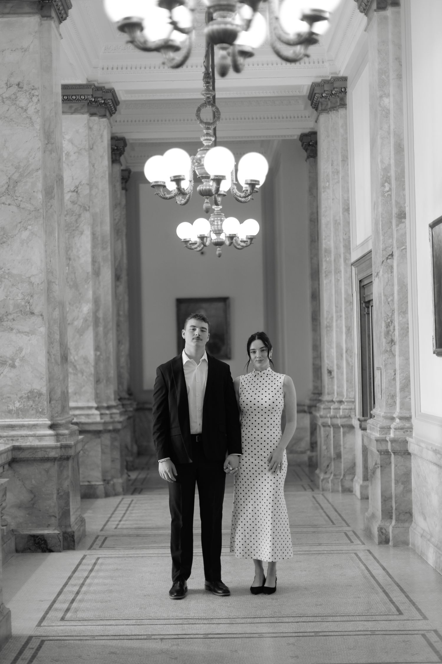 Black and white photo of couple standing in elegant marble hallway with chandelier overhead.