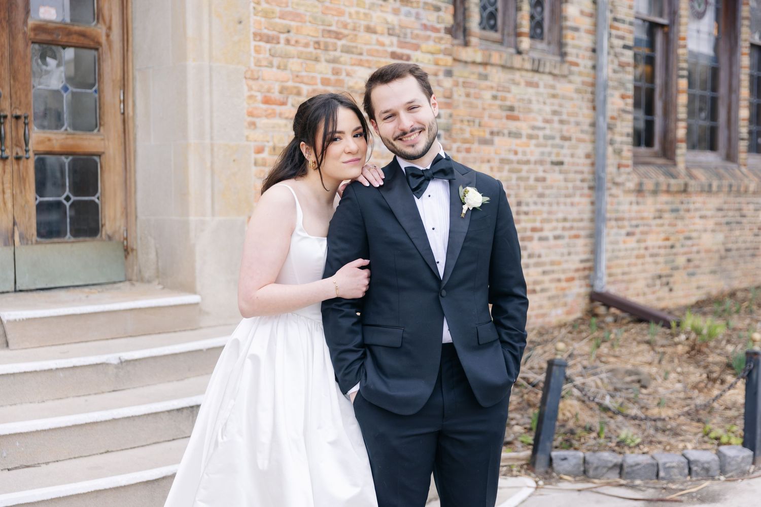 Bride in white gown and groom in black tuxedo posing together outside a brick building on their wedding day.