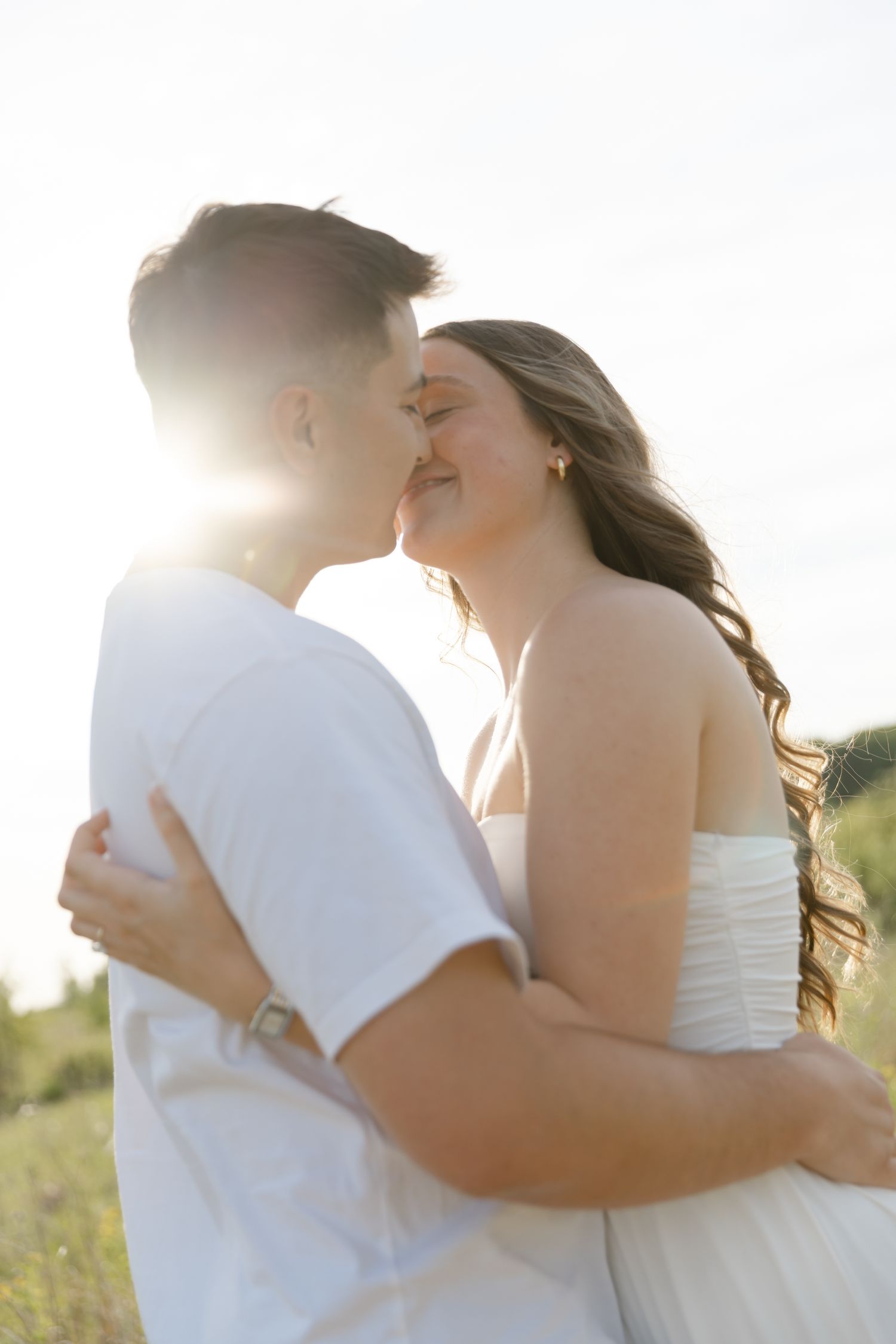Couple sharing a romantic kiss outdoors in golden sunlight, woman in white dress embracing man in white t-shirt.