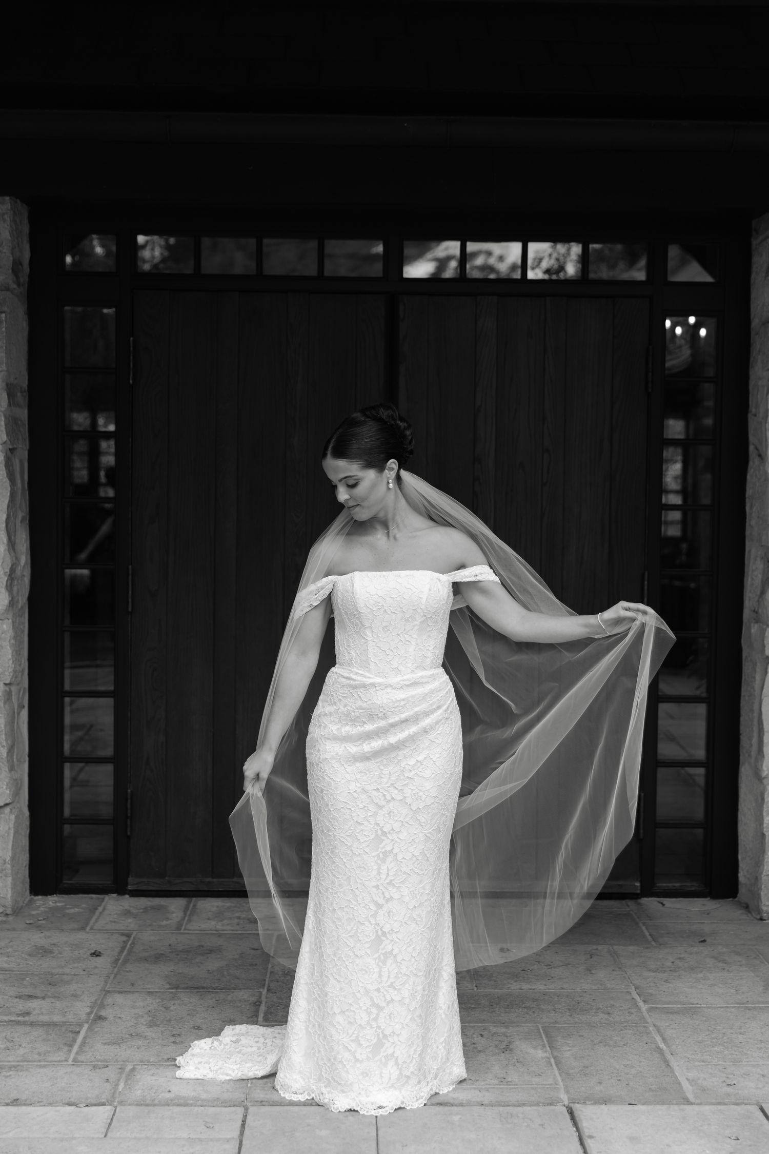 Bride in off-shoulder lace wedding gown holding flowing veil, standing before dark wooden doors in black and white photo.
