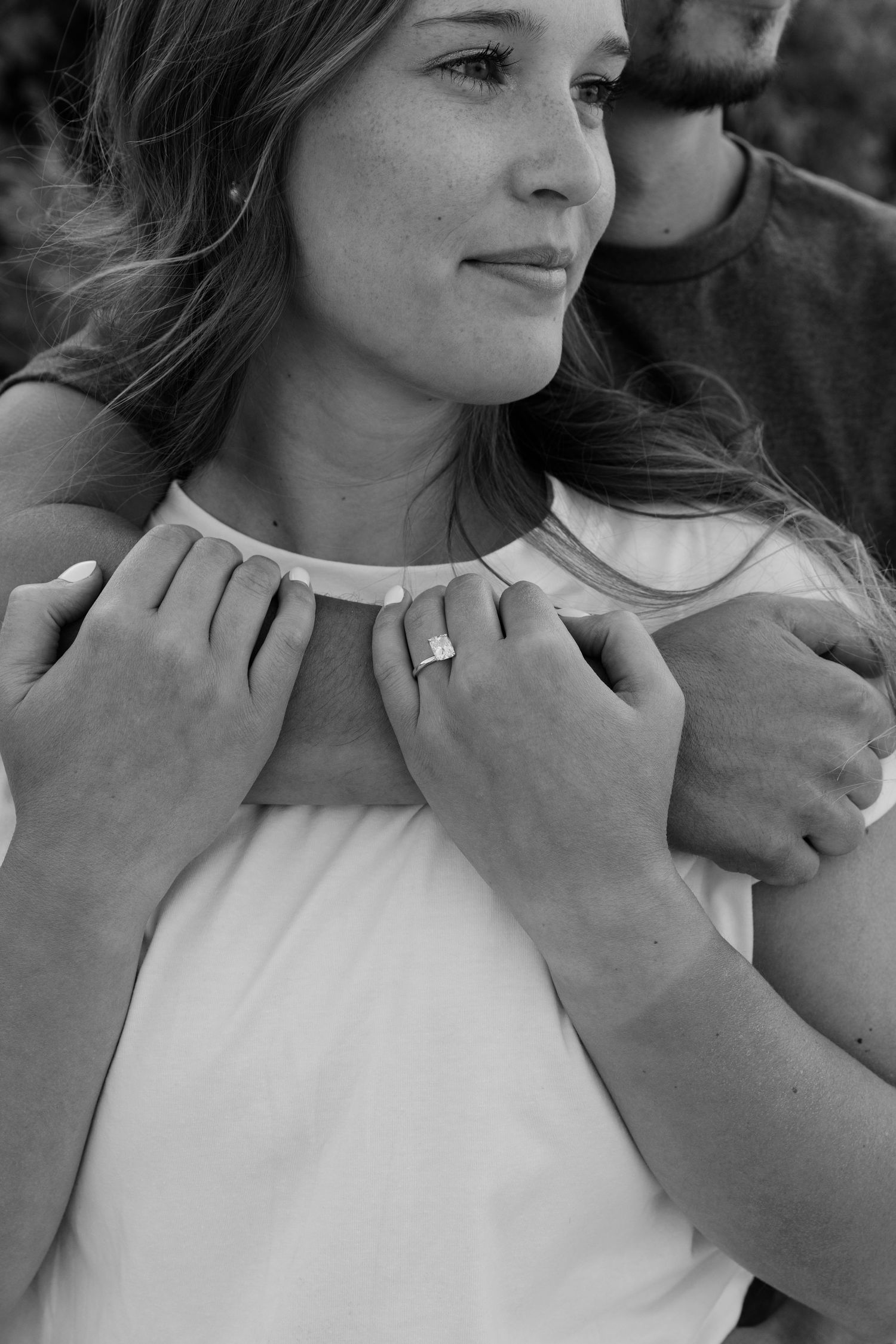 Black and white photo of a couple embracing, woman wearing engagement ring and necklace, man's hands on her shoulders.