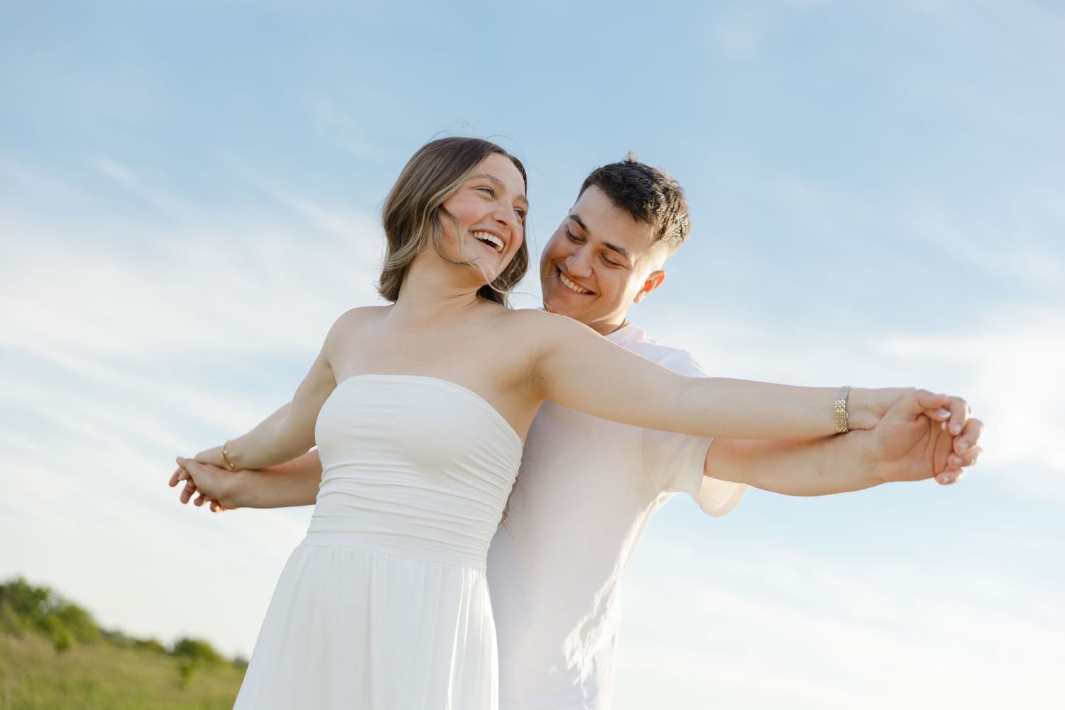 Happy couple in white dancing outdoors under blue sky, woman laughing joyfully as man embraces her from behind.
