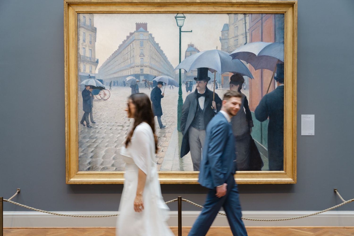 Couple walking past Gustave Caillebotte's Paris Street Rainy Day painting in a museum gallery.
