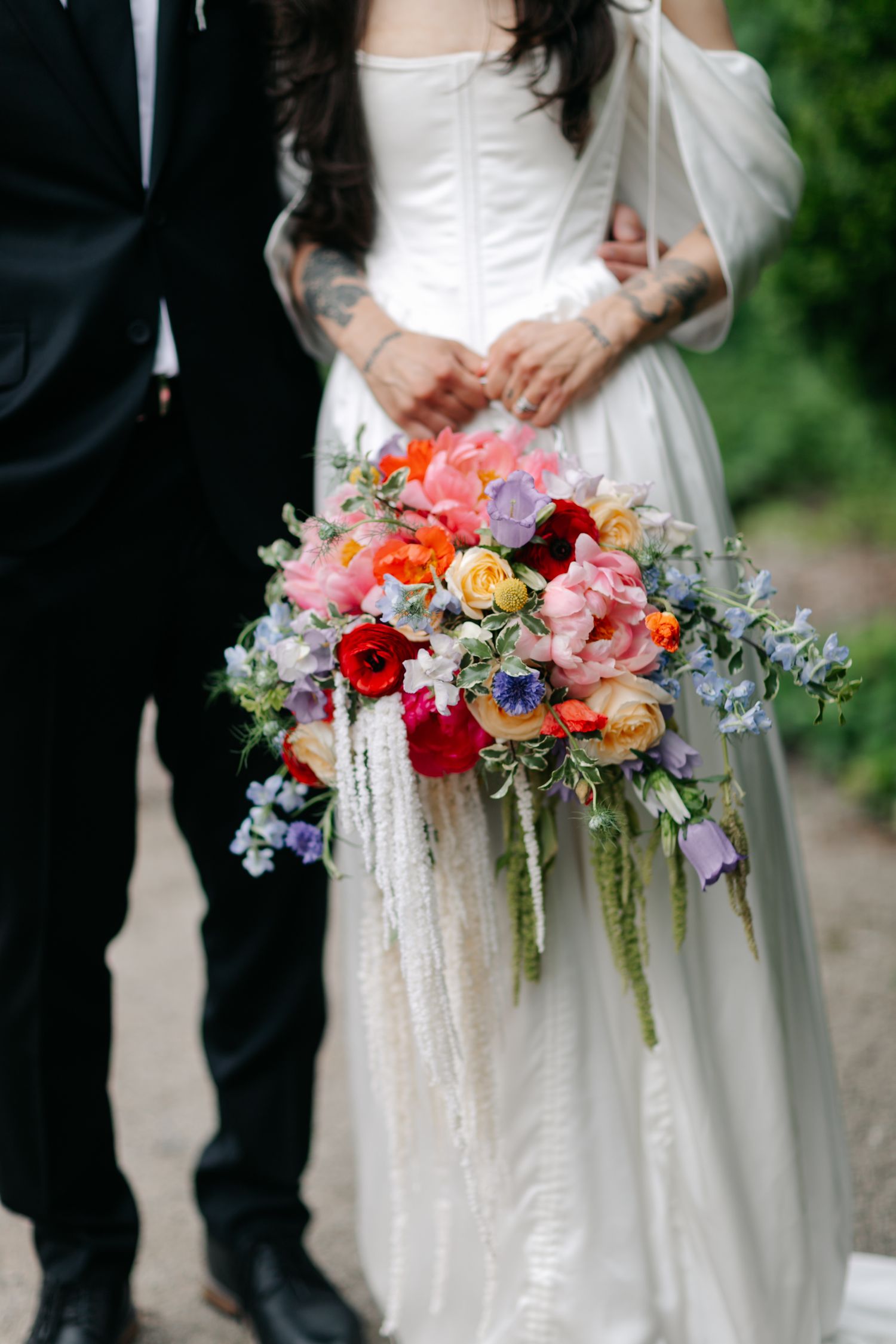 Bride holding a vibrant cascading wedding bouquet with red, pink, and blue flowers beside groom in black suit.
