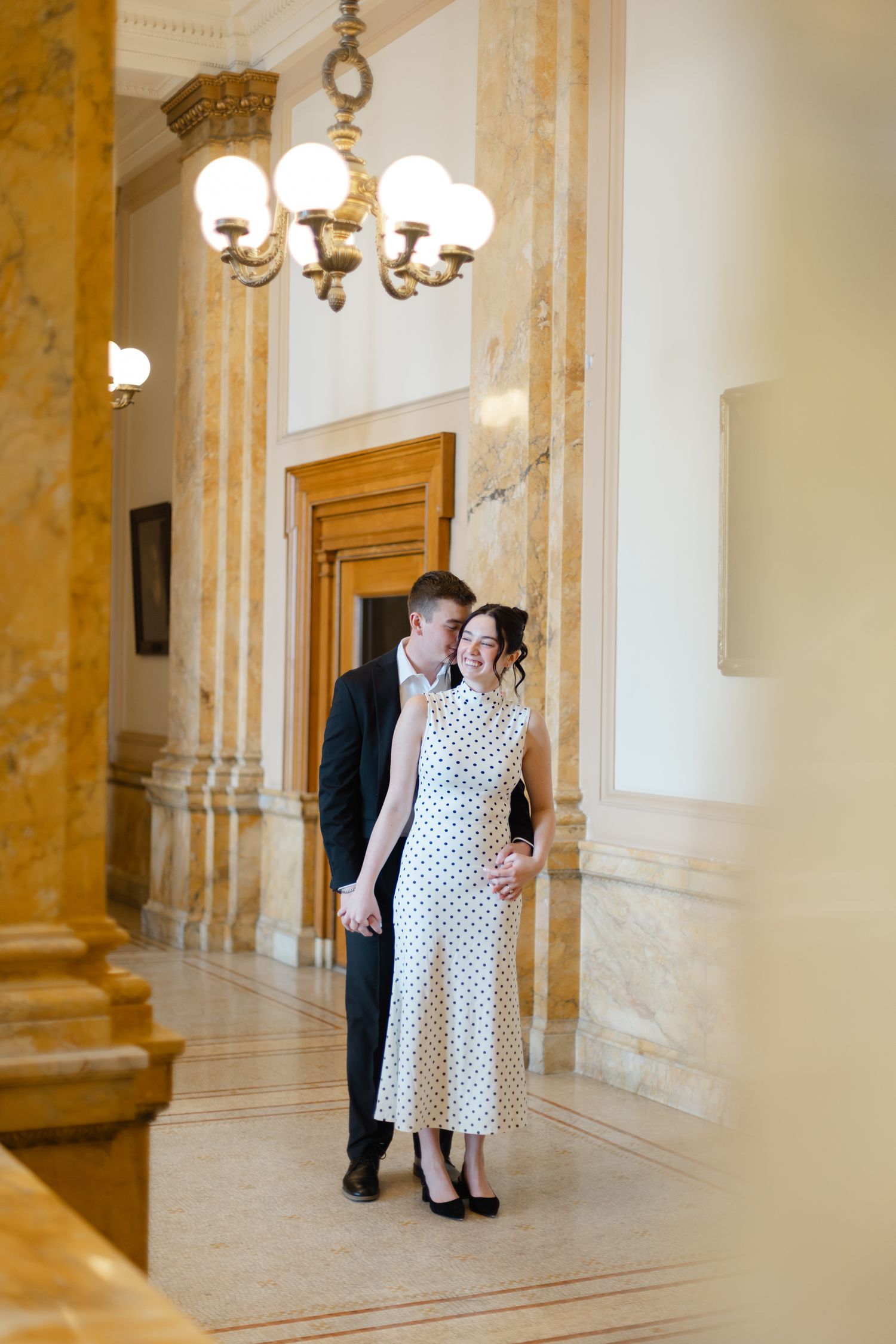 Couple embracing in elegant marble hallway, woman in polka dot dress, man in dark suit, chandelier overhead.