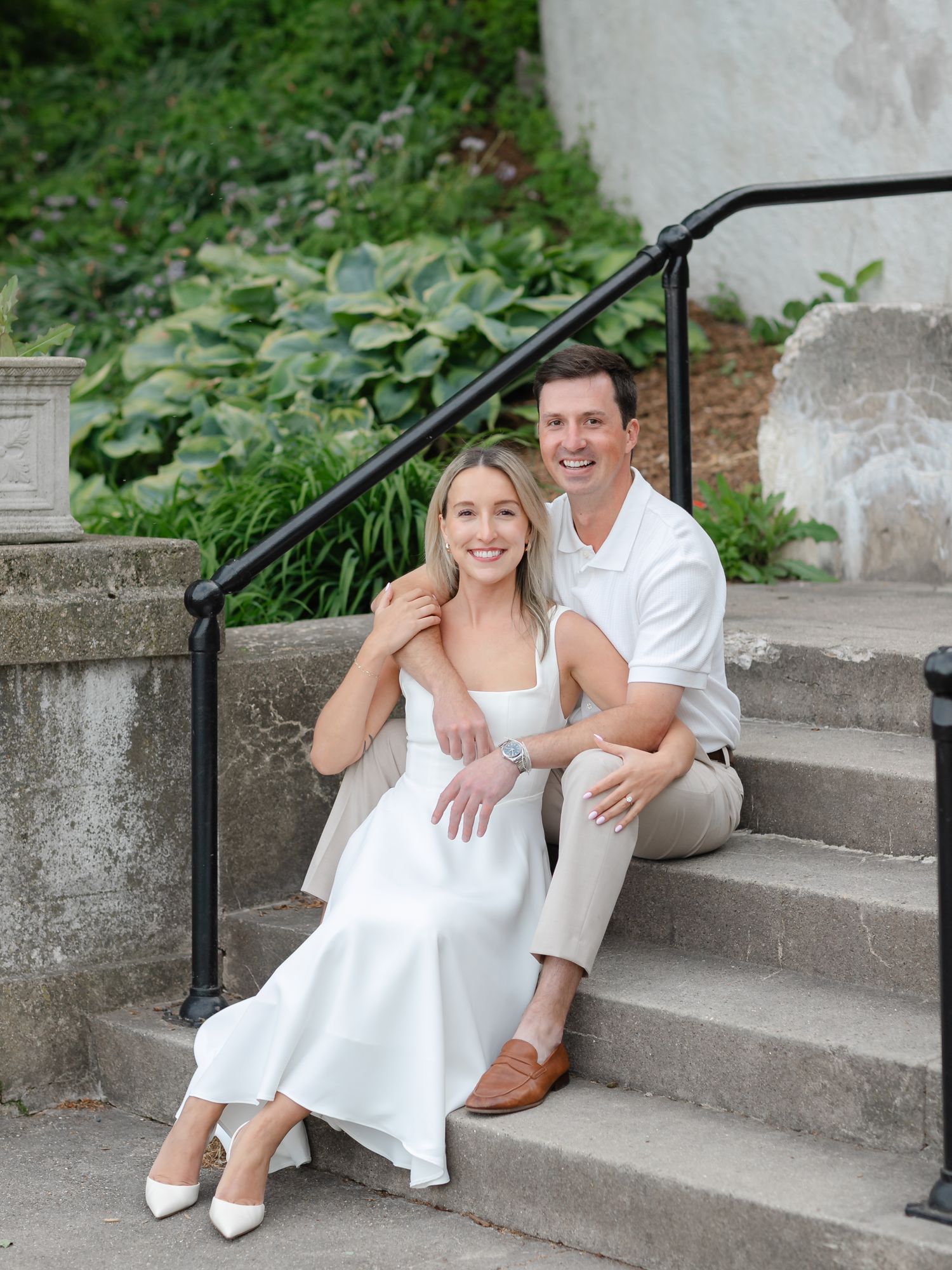 A smiling couple poses on stone steps, woman in white dress and heels, man in white shirt and tan pants, lush greenery behind.