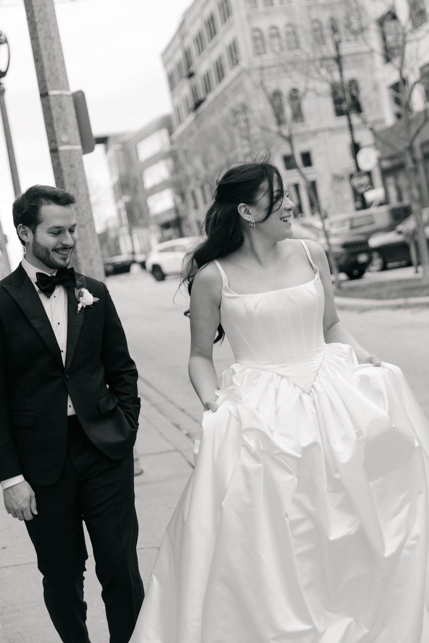Black and white photo of a smiling bride and groom walking together on a city street, she in a full gown, he in a tuxedo.