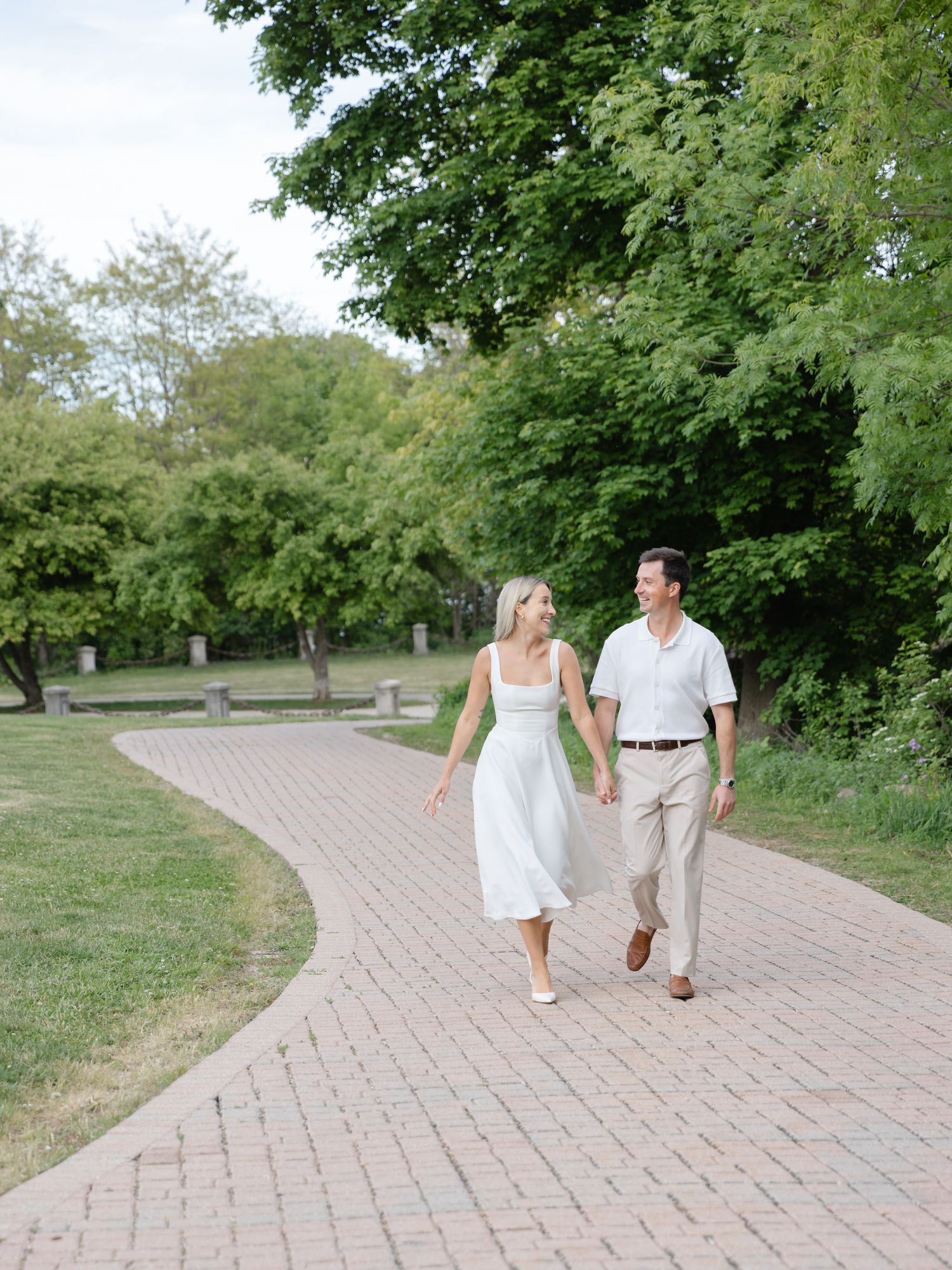 A couple holds hands and smiles while walking along a brick path surrounded by lush green trees.