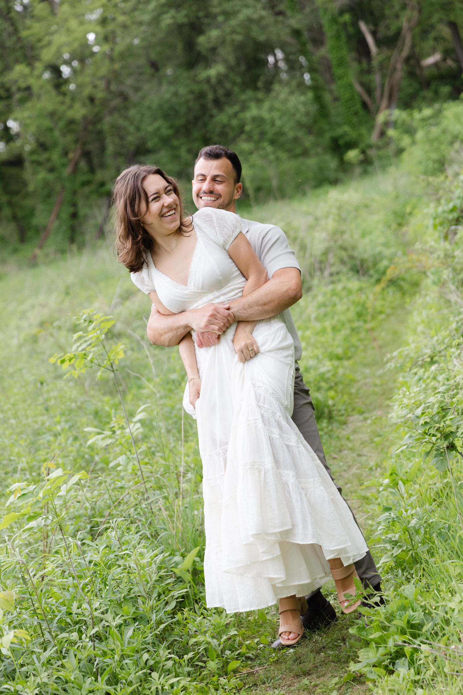 A joyful couple embraces and laughs together in a lush green outdoor setting, the woman wearing a flowing white dress.