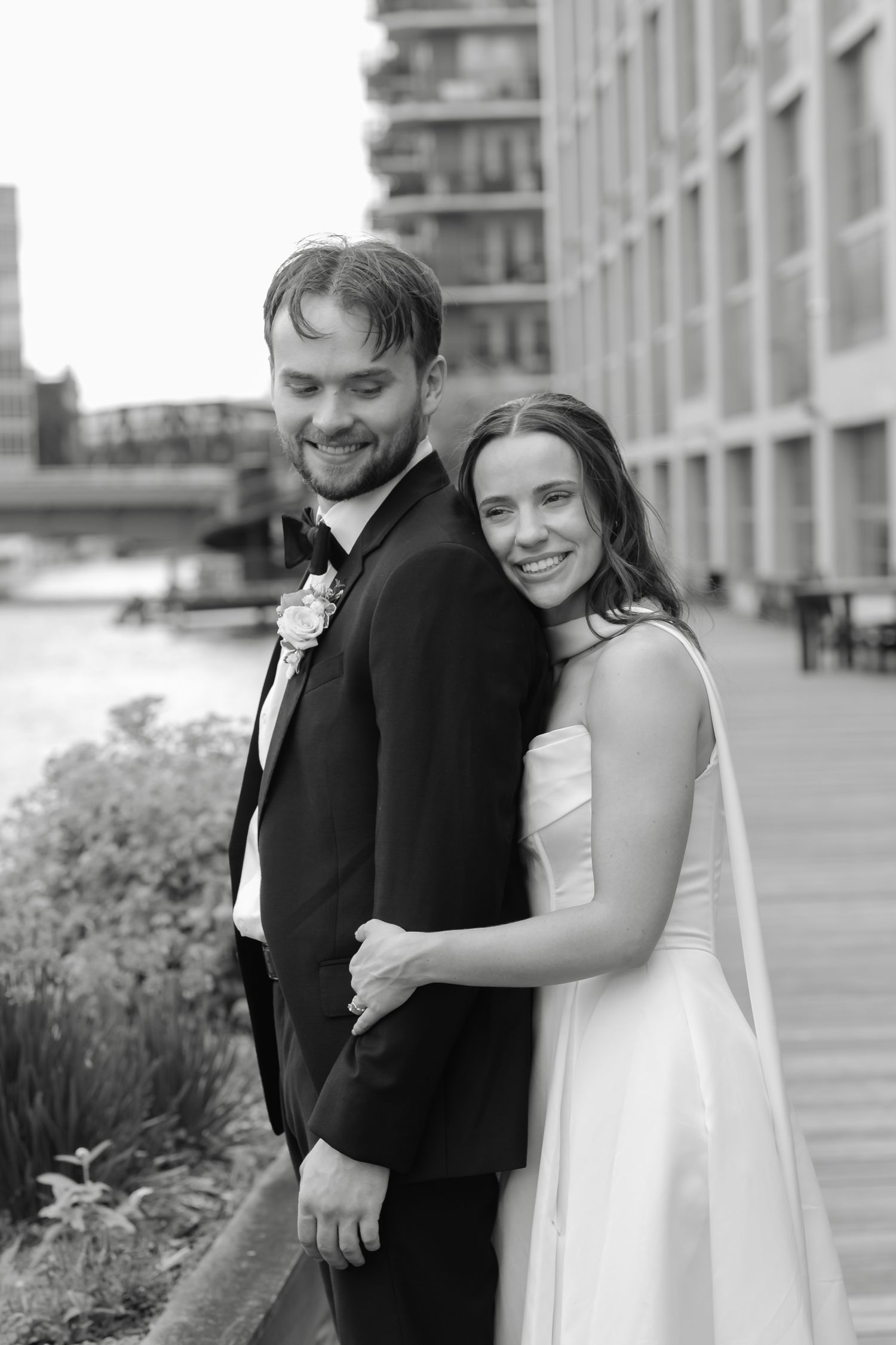 Black and white wedding portrait of smiling bride and groom posing together outdoors near a waterfront.