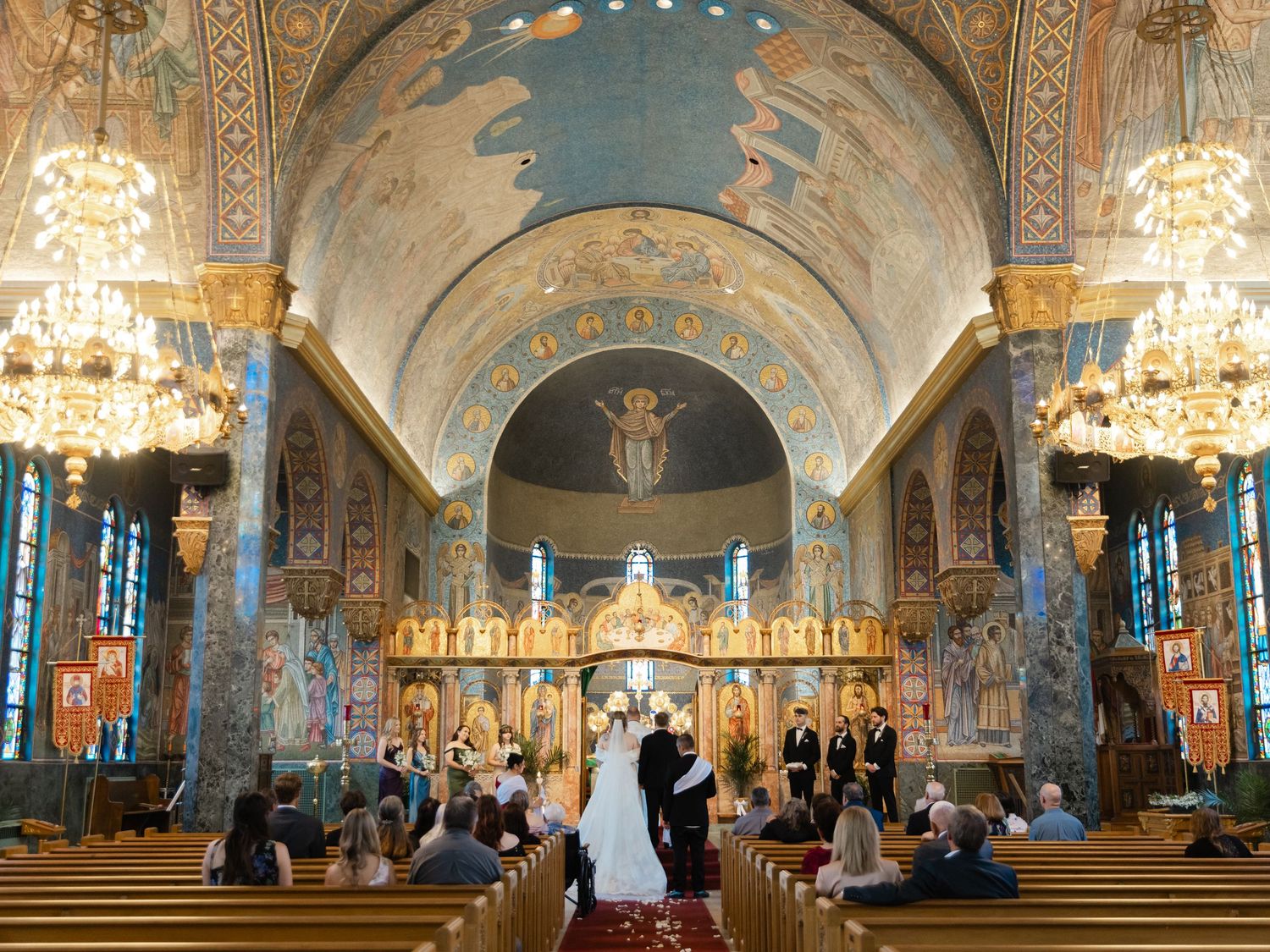 Wedding ceremony in ornate Orthodox church with stunning frescoes, golden iconostasis, and elegant chandeliers.