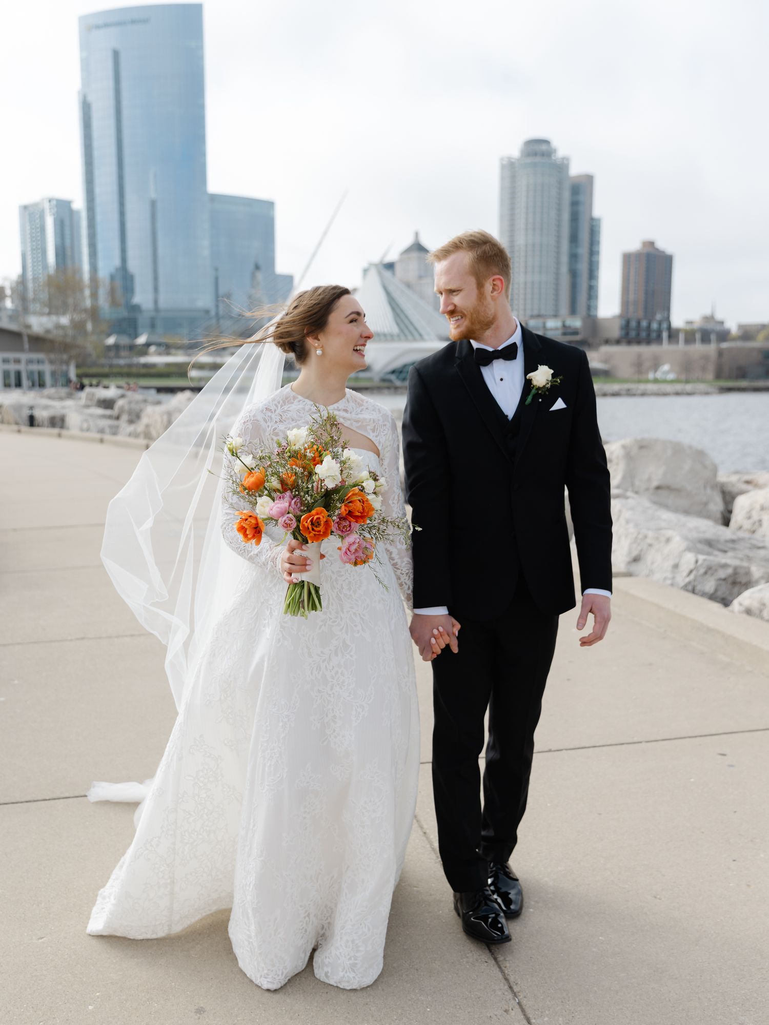 Smiling bride and groom walk hand in hand along waterfront path with veil flowing and city skyline behind them.