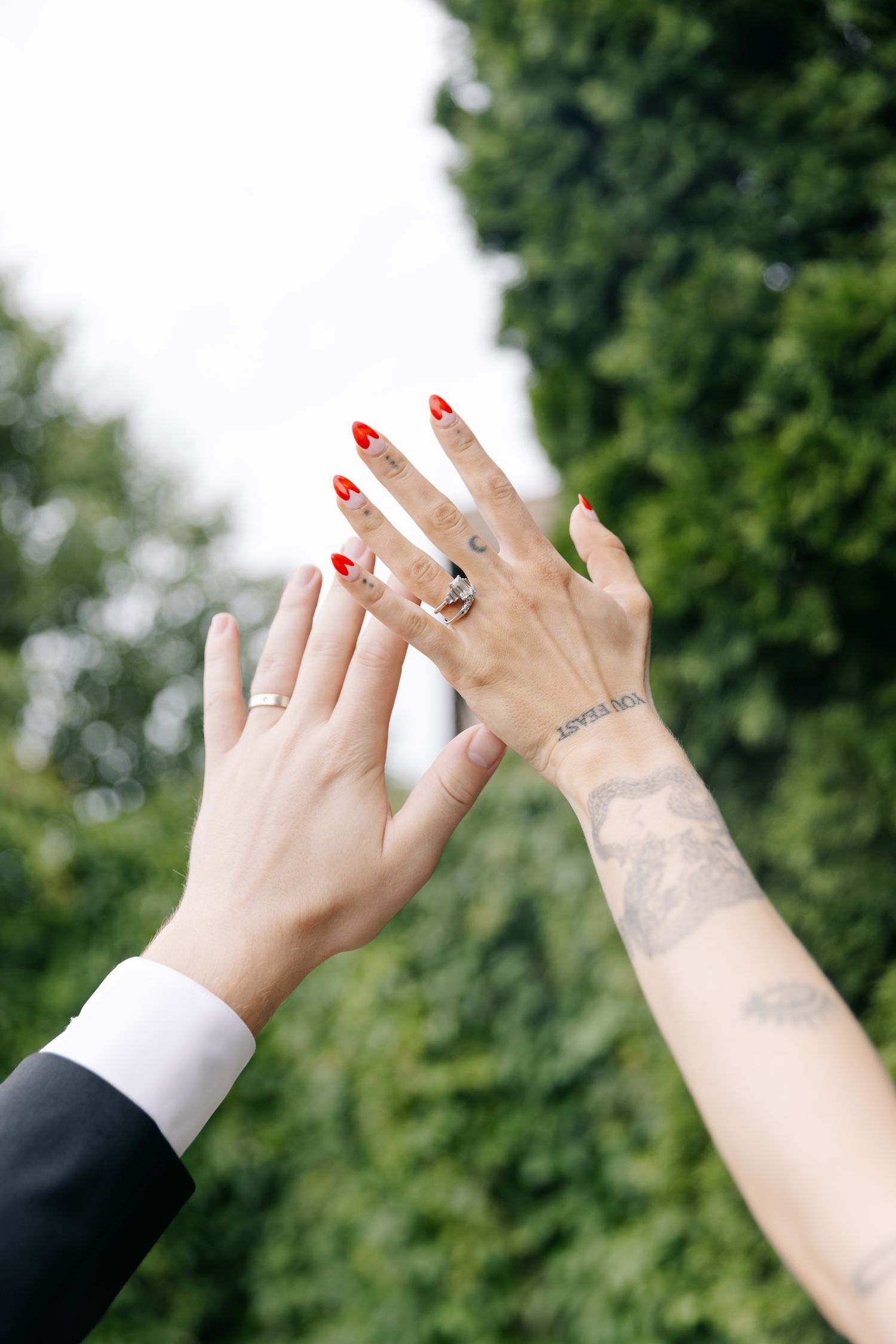 Bride and groom displaying wedding rings, bride with red nails and tattoos raising hands against green trees.